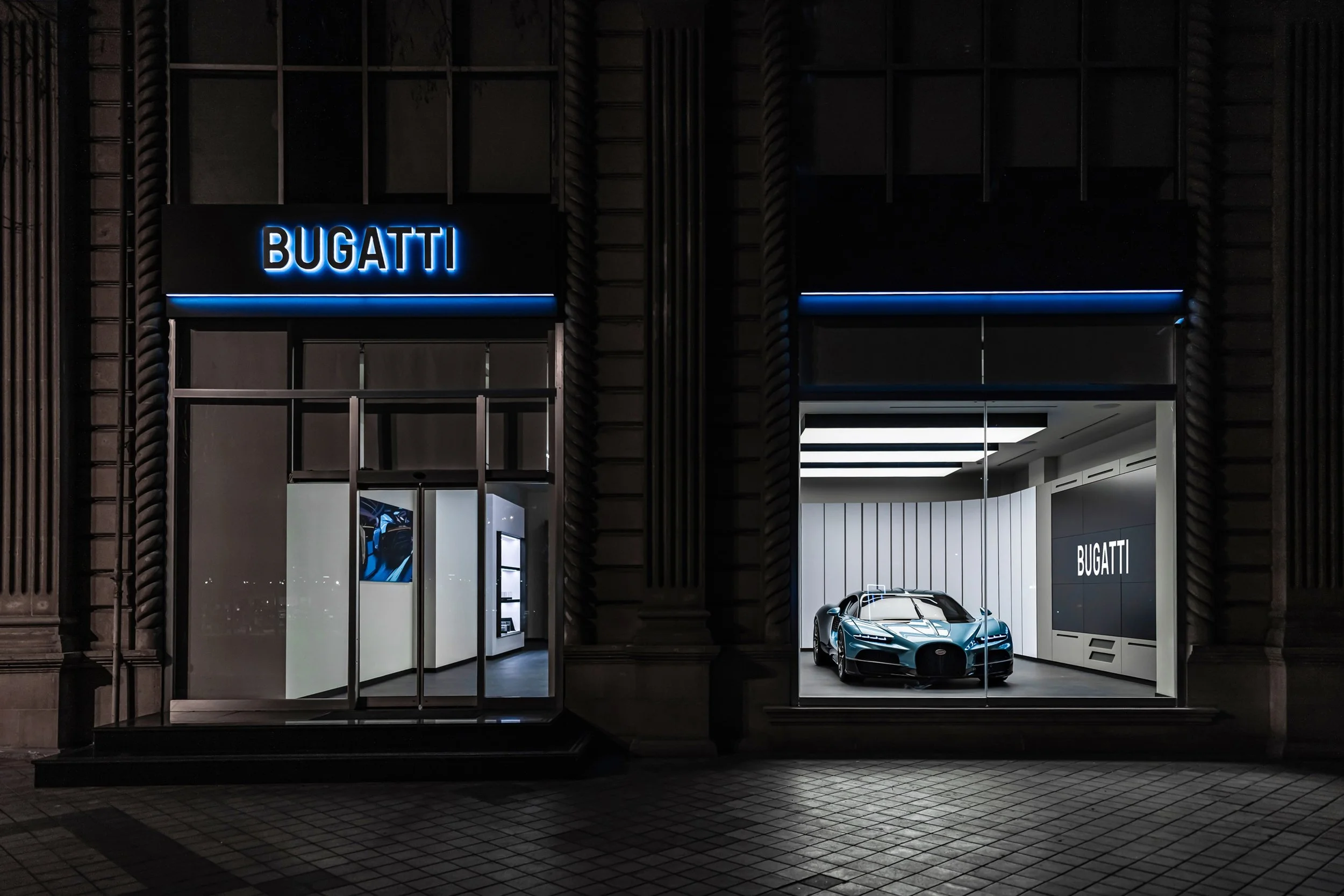 Nighttime view of a Bugatti showroom with a single blue Bugatti car on display in a well-lit interior, seen through large glass windows, with the Bugatti sign illuminated above the entrance.