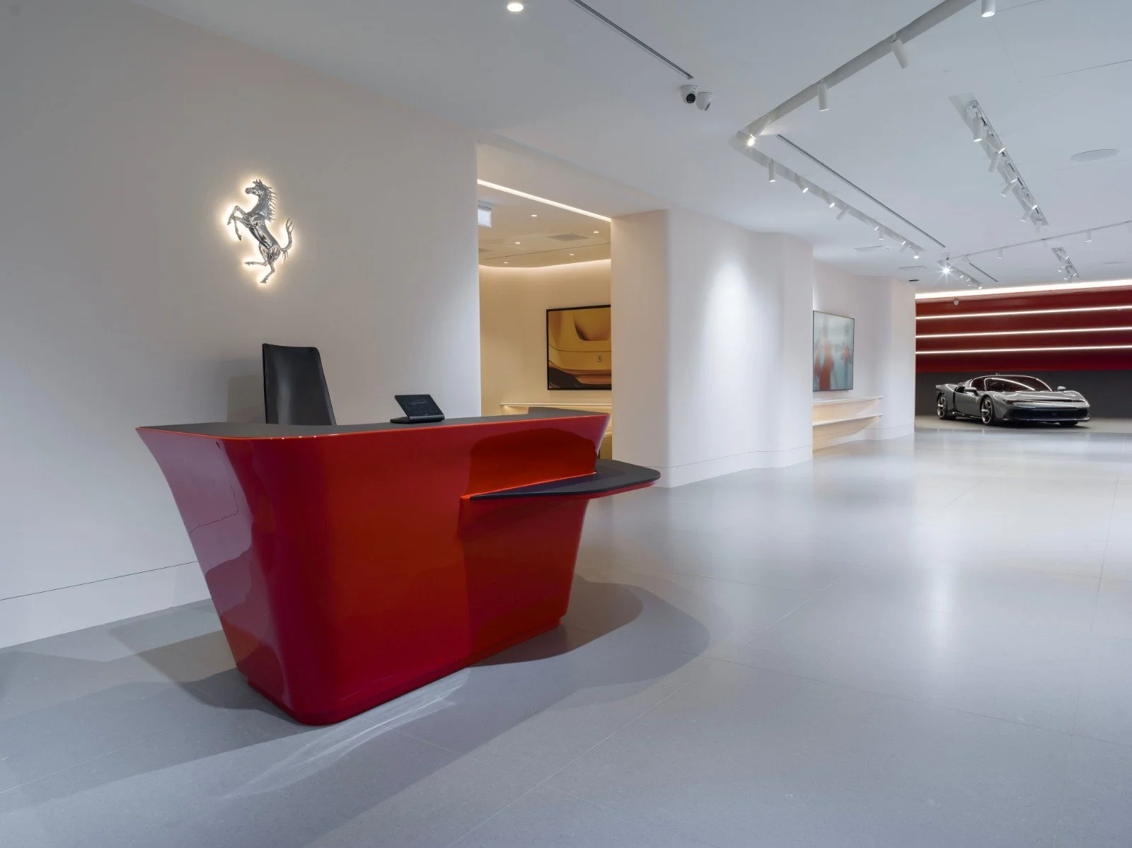 Luxury car showroom with a red reception desk, a Ferrari logo on the wall, and a sleek silver sports car in the background.