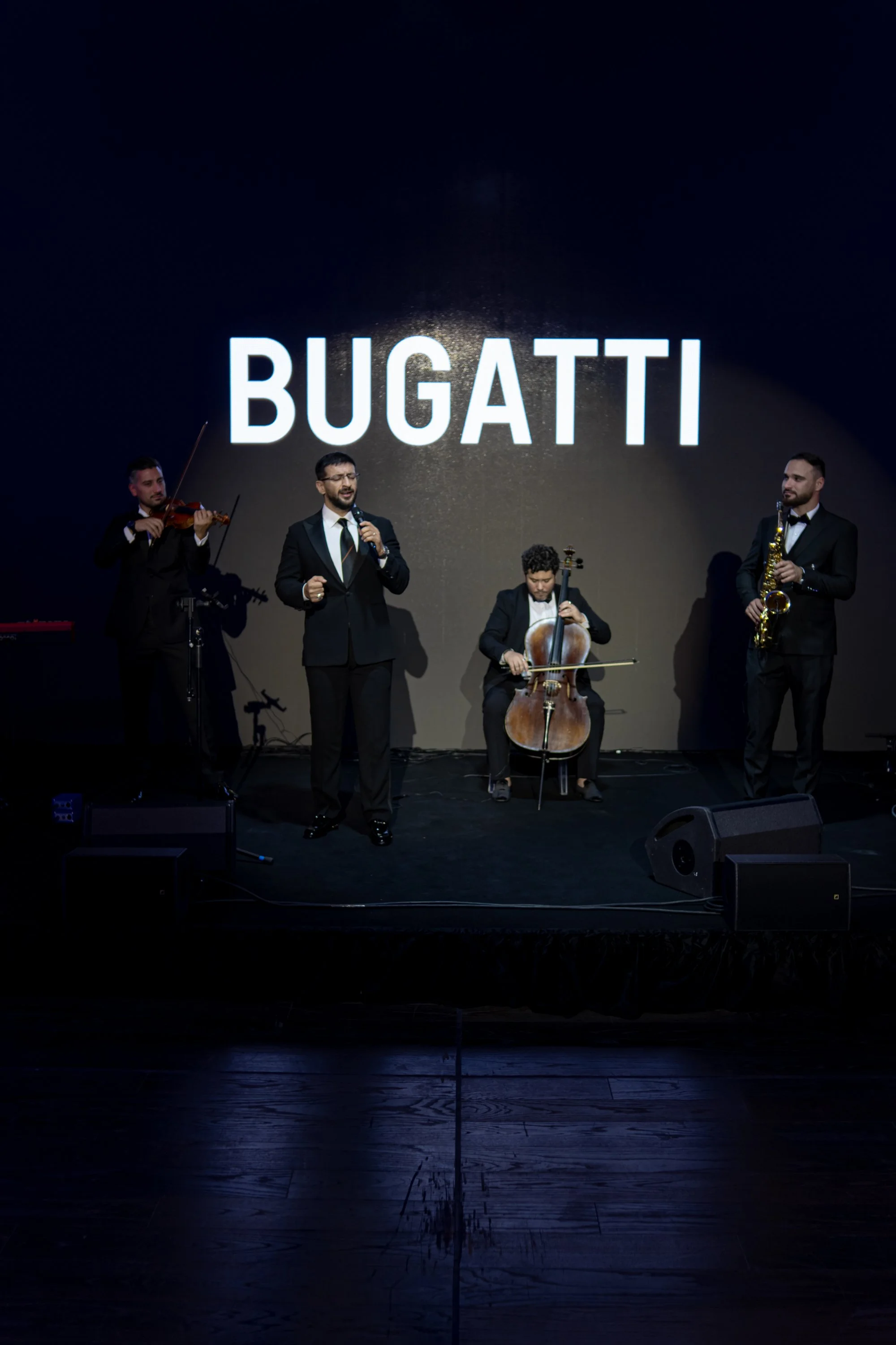 A musical performance with four musicians in formal attire on stage, with a large screen behind them displaying the word 'BUGATTI'. One musician is singing, and three are playing instruments: violin, cello, and saxophone.