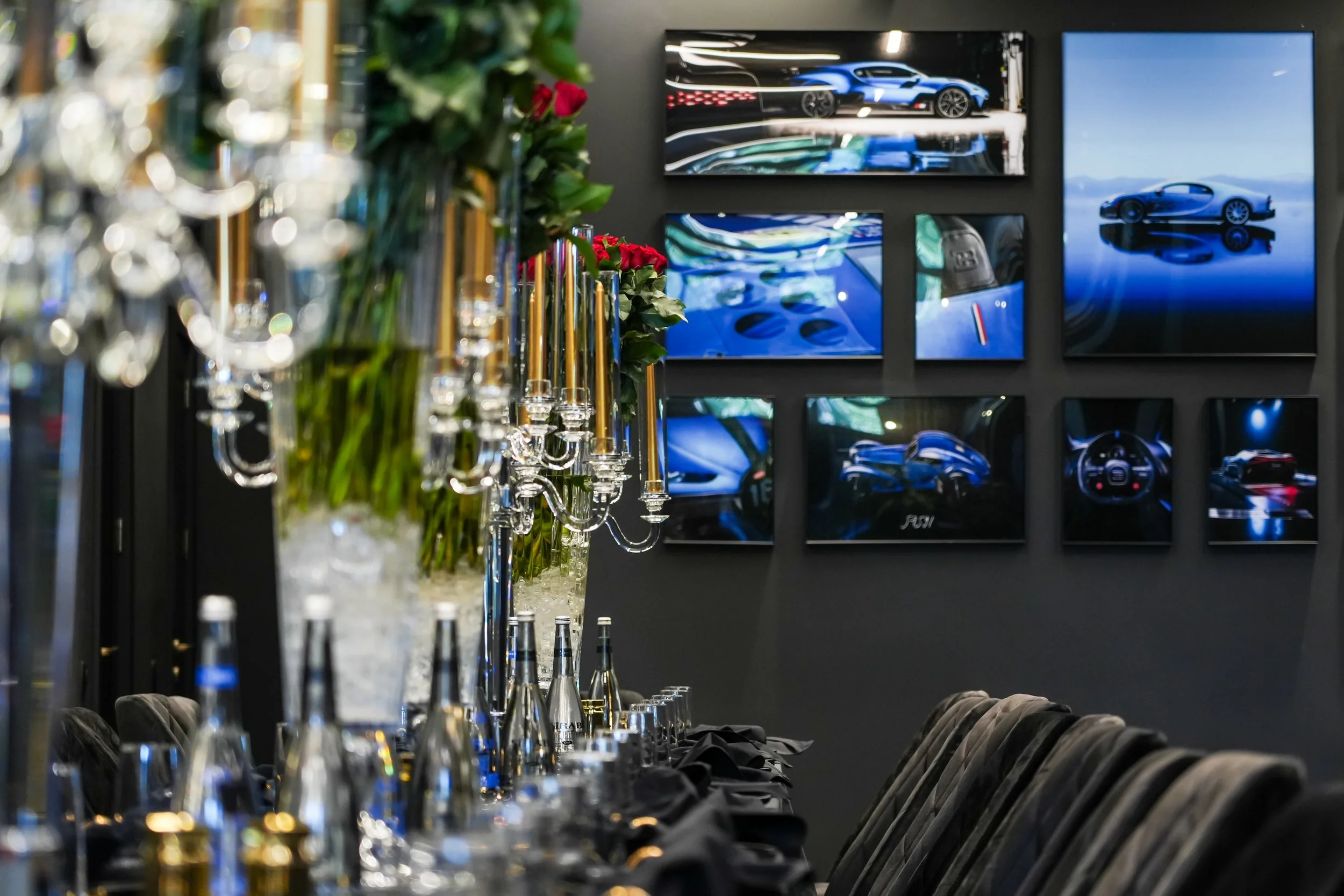 Elegant dining setup with tall floral arrangements, candelabras, and tableware, featuring a wall decorated with multiple screens displaying luxury cars in a sleek showroom or event setting.