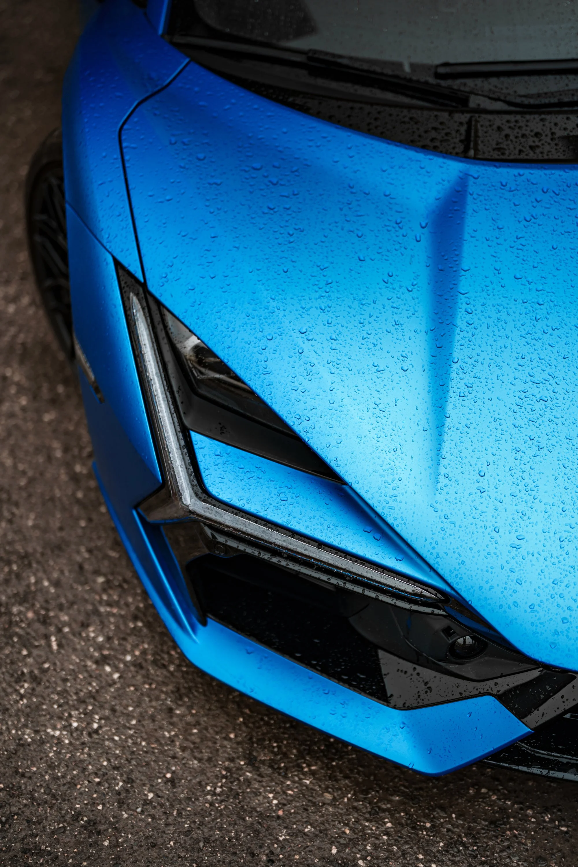 Close-up of the front right side of a blue car with water droplets on its surface, on a wet asphalt surface.