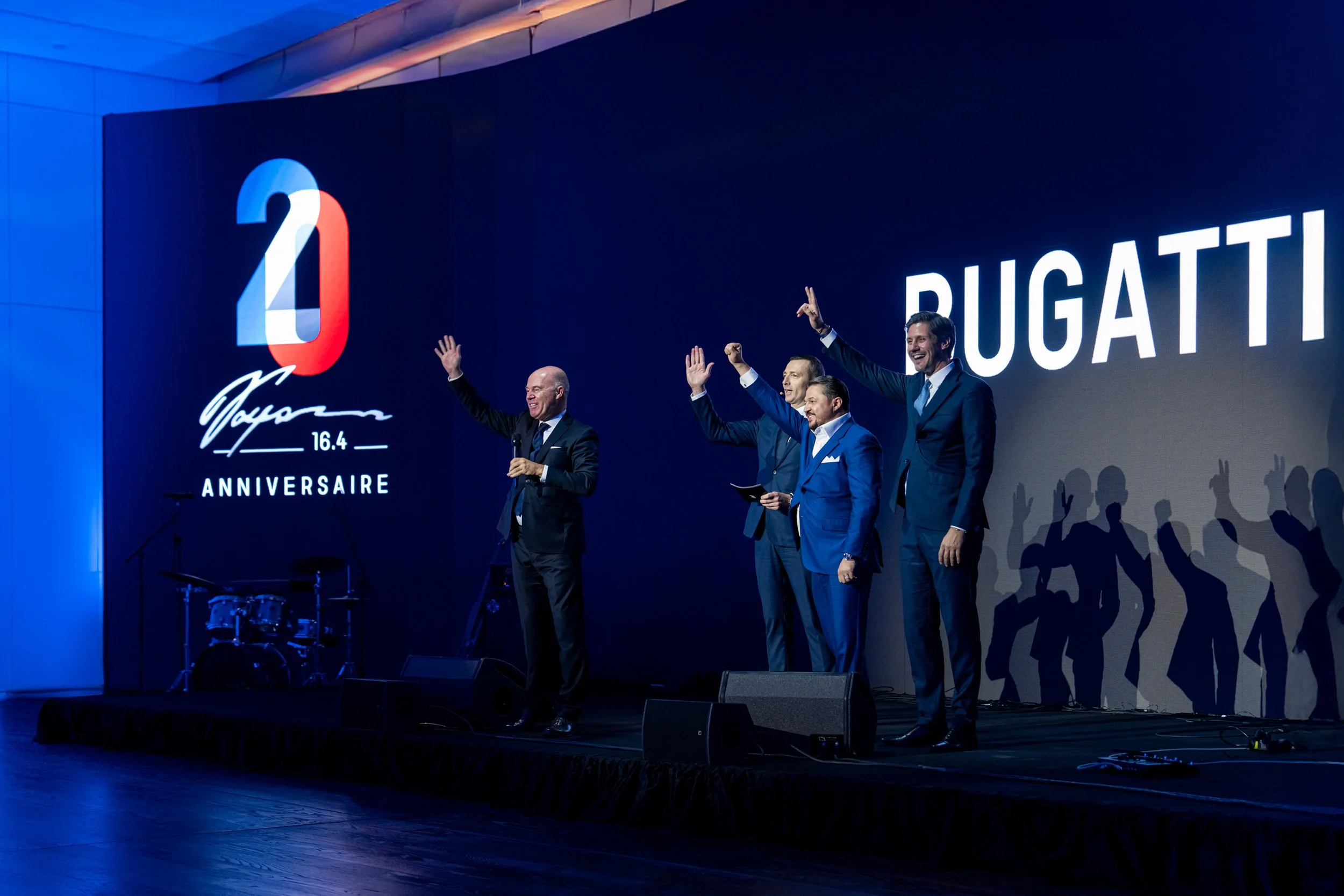 Four men in suits on stage celebrating at a Bugatti event with a large screen displaying the Bugatti logo, the number 16.4, and the word 'Anniversaire'.