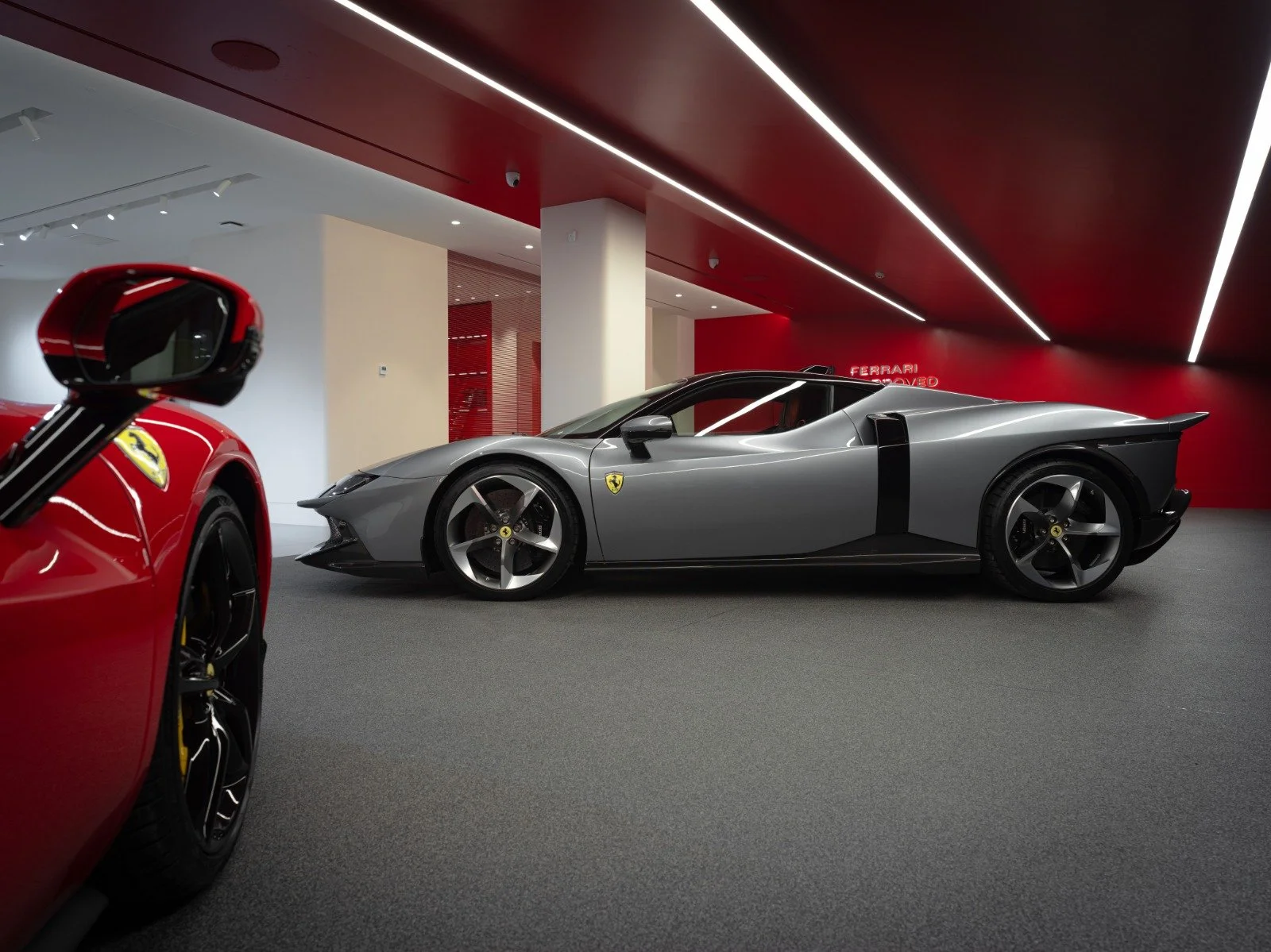 A modern car showroom displaying a silver Ferrari sports car with sharp, aerodynamic design and large rear wheel, with part of a red Ferrari visible in the foreground. The background features a red wall with the words "Ferrari Approved".