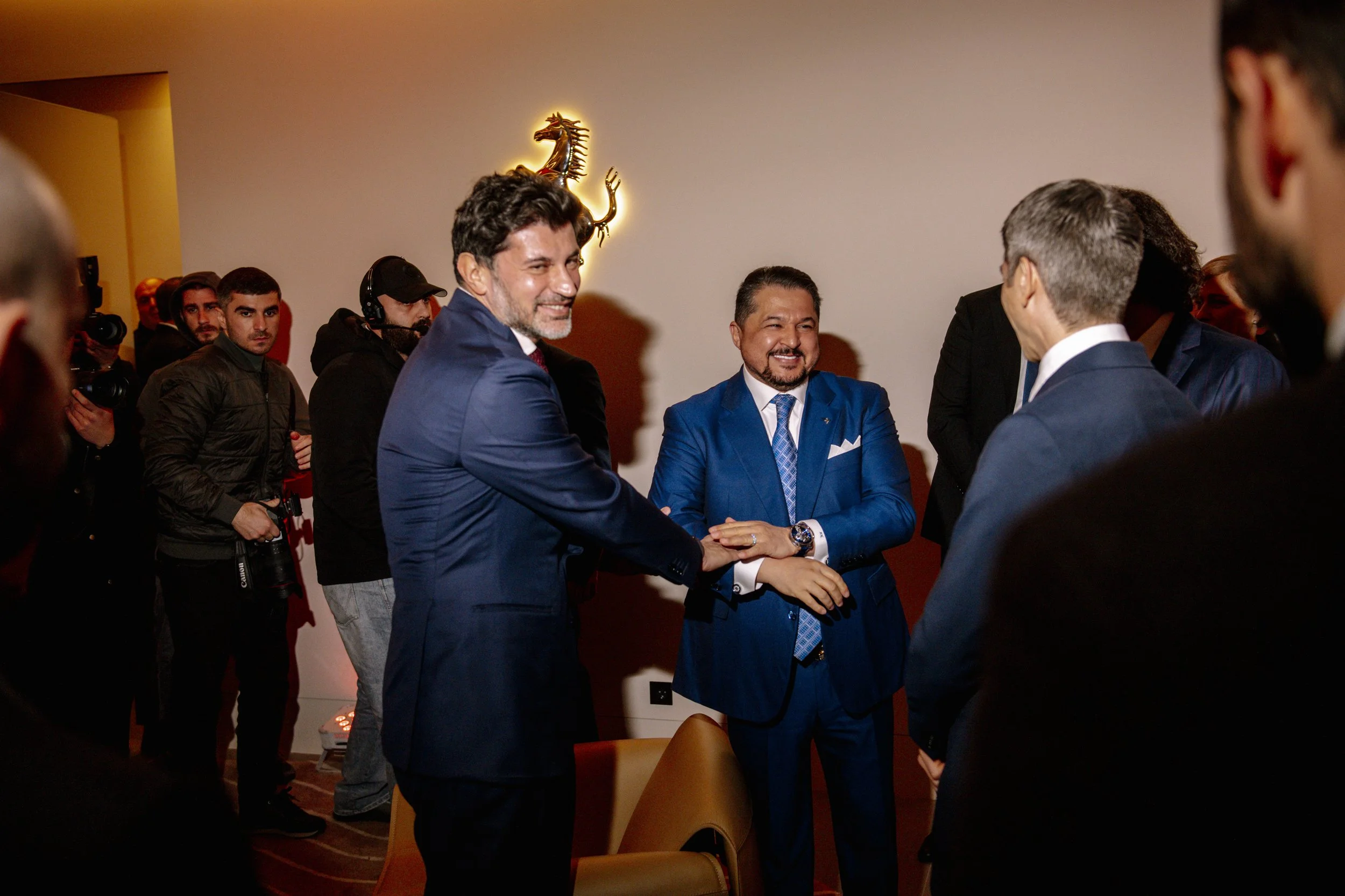 Men in suits shaking hands and smiling at an indoor event with a dragon emblem on the wall behind them, surrounded by other people, some taking photos.
