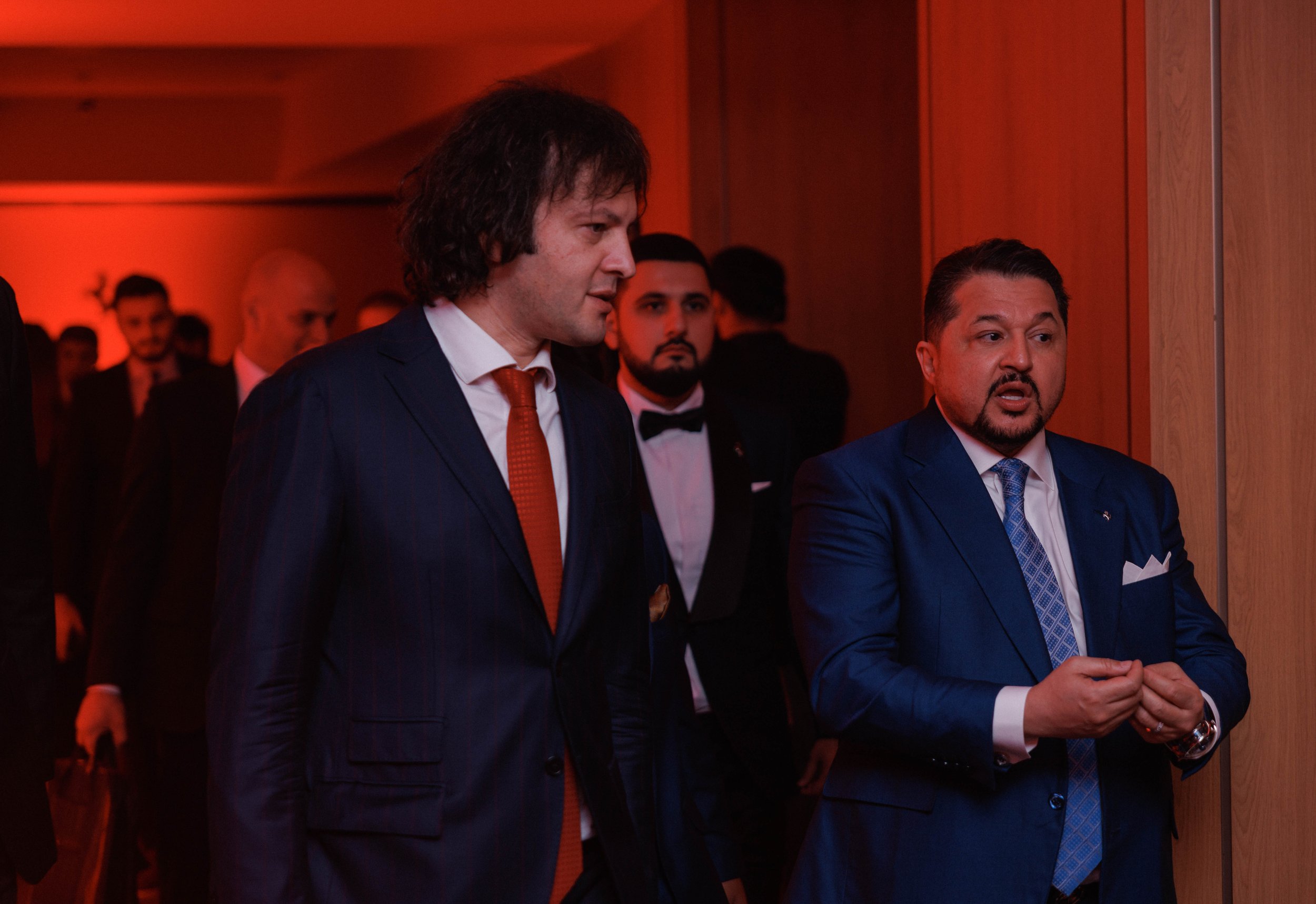 Two men in suits engaging in conversation at a formal event, with several other formally dressed individuals in the background.