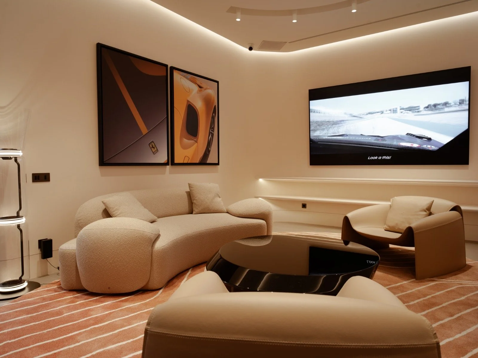 A modern living room with beige and cream furniture, including a curved sofa, a single armchair, and a black coffee table. There are three framed pictures on the wall, one of which features a yellow sports car. A large flat-screen TV displays a racing scene with the caption "Look at this!" The room has a soft beige and cream color scheme with subtle lighting accents.