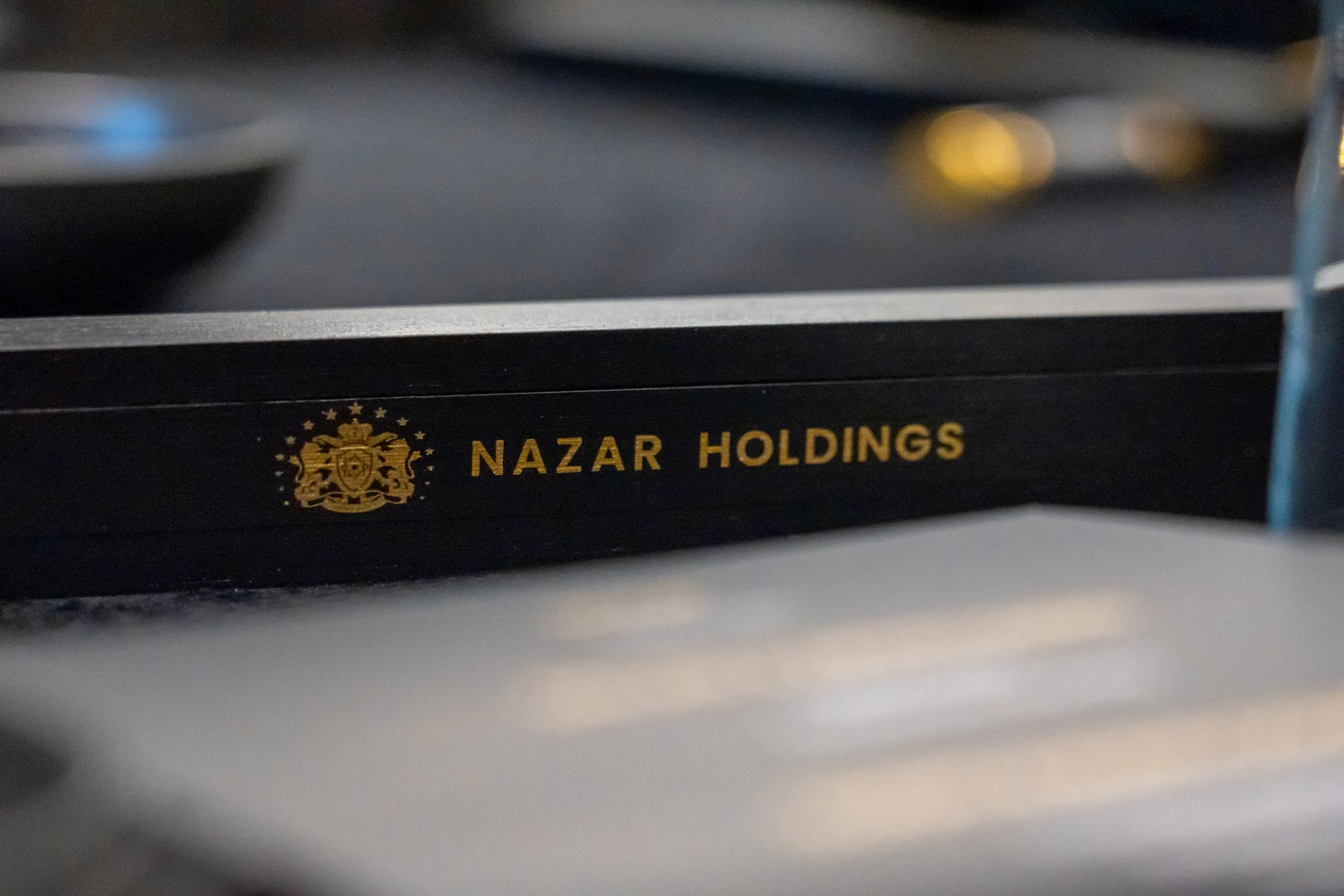 Close-up of a black tray with gold text reading 'NAZAR HOLDINGS' and a gold crest, partially visible, on a dark table surface.
