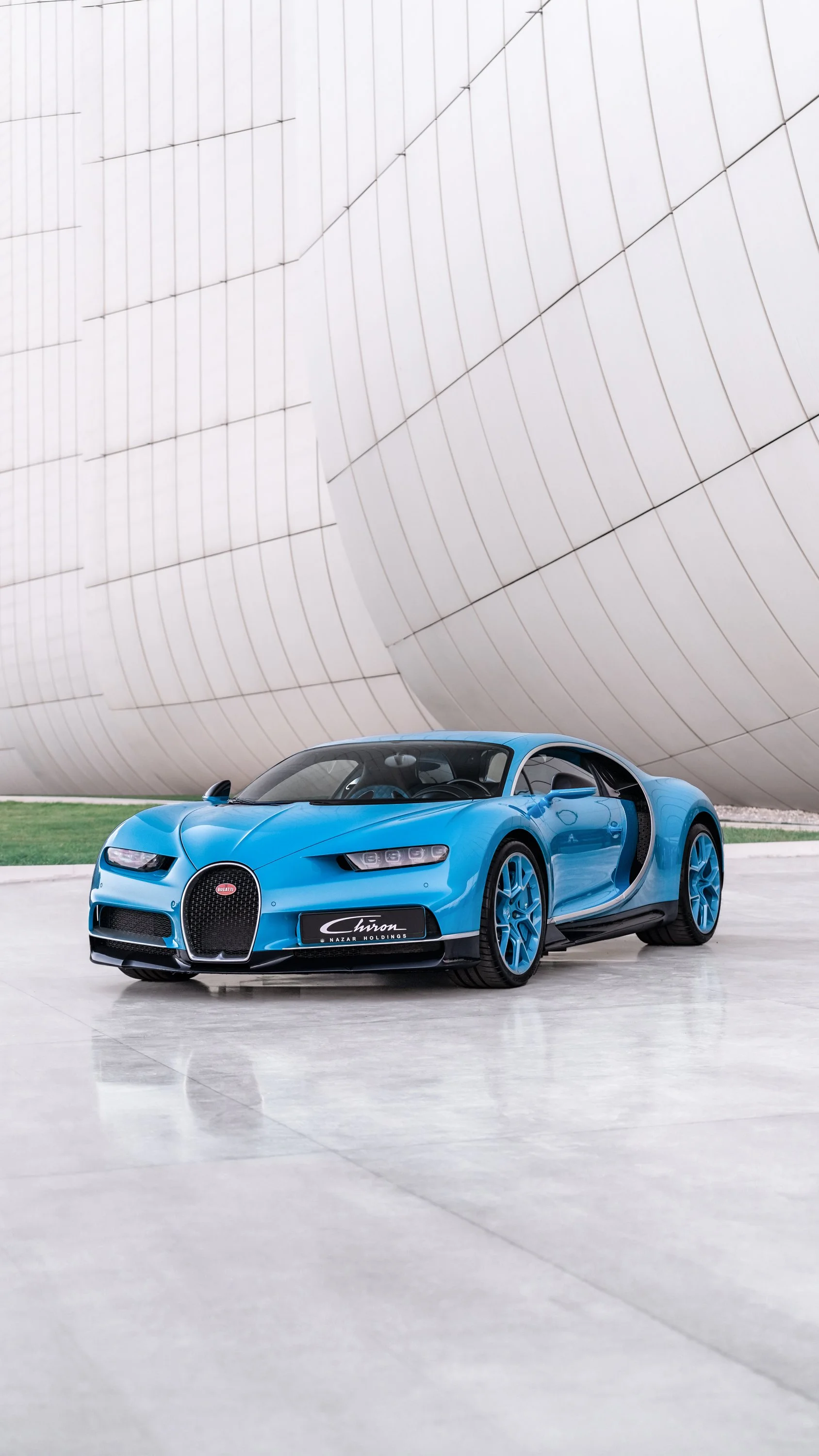 Blue Bugatti Chiron sports car parked on a modern concrete surface in front of a curved architectural structure.