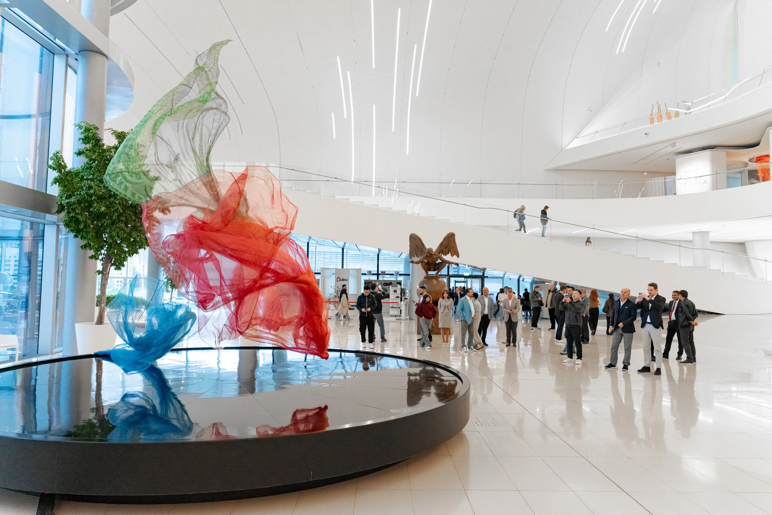 Interior of a modern building with a large colorful fabric sculpture in the foreground, and people walking and gathering in the background.