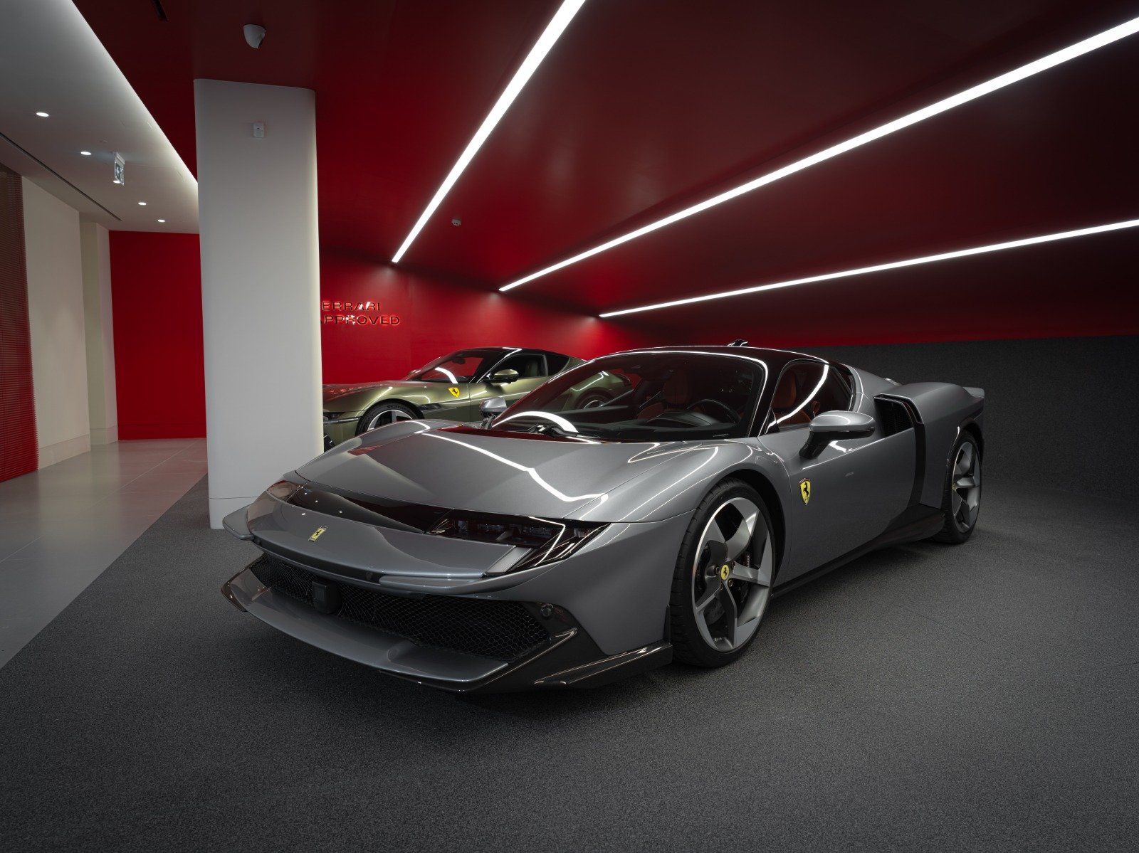 A sleek silver Ferrari sports car with black accents and modern design in a luxury showroom, with another Ferrari in the background. Red walls and ceiling with stylish lighting.