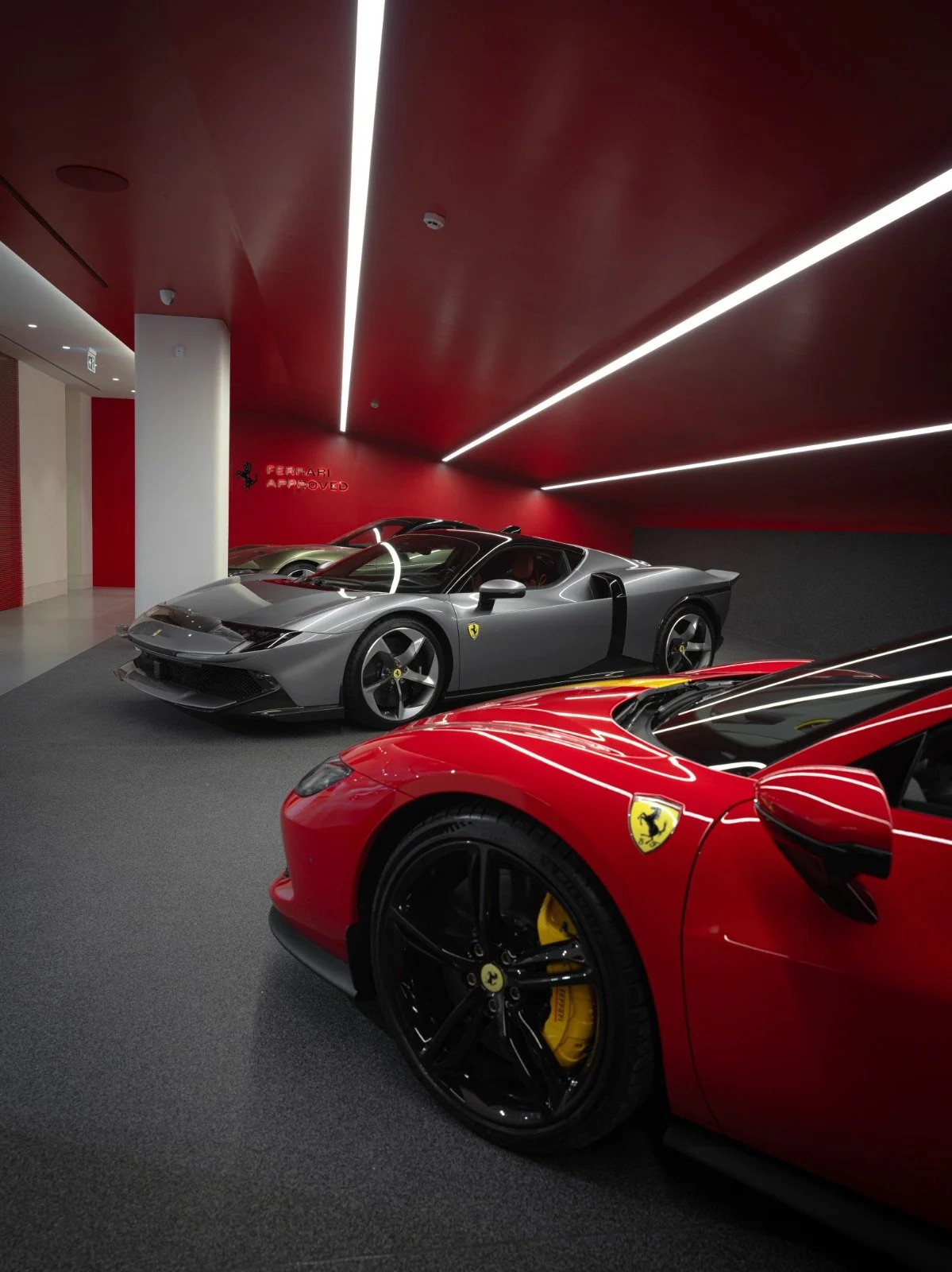 Three luxury sports cars displayed in a showroom with red and black walls. The front car is red with black wheels and yellow brake calipers, the middle car is gray, and the back car is a darker shade with a sleek design.