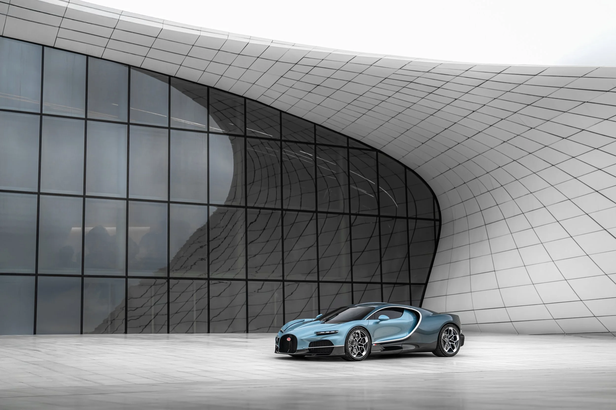 A sleek, blue Bugatti sports car parked in front of a modern architectural building with large glass windows and curved, white panels.