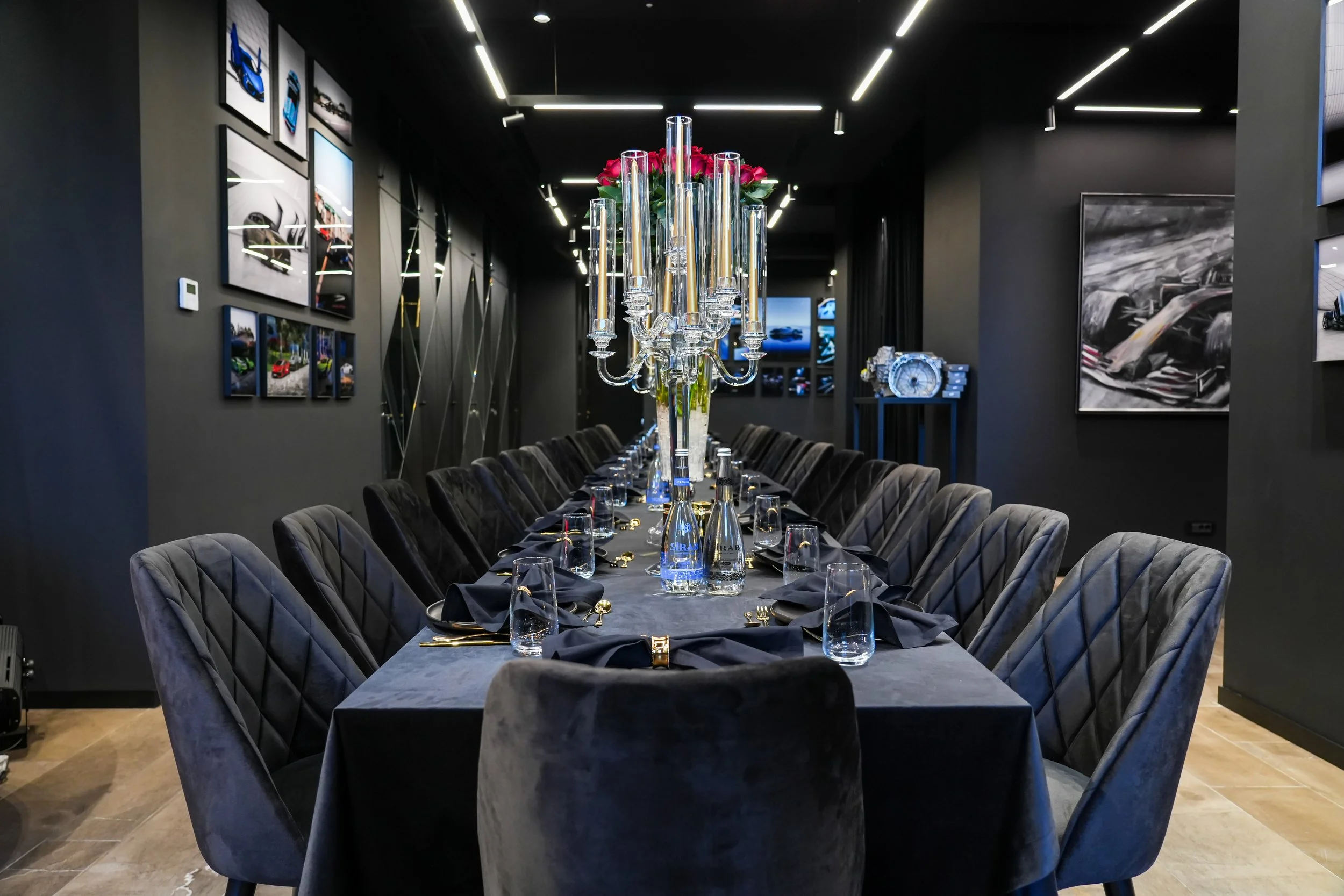A luxurious dining room set up with a long black table, surrounded by black velvet chairs, with a centerpiece of tall glass candelabras and flowers. The walls are decorated with automotive-themed artwork, and the ceiling features sleek black and whit