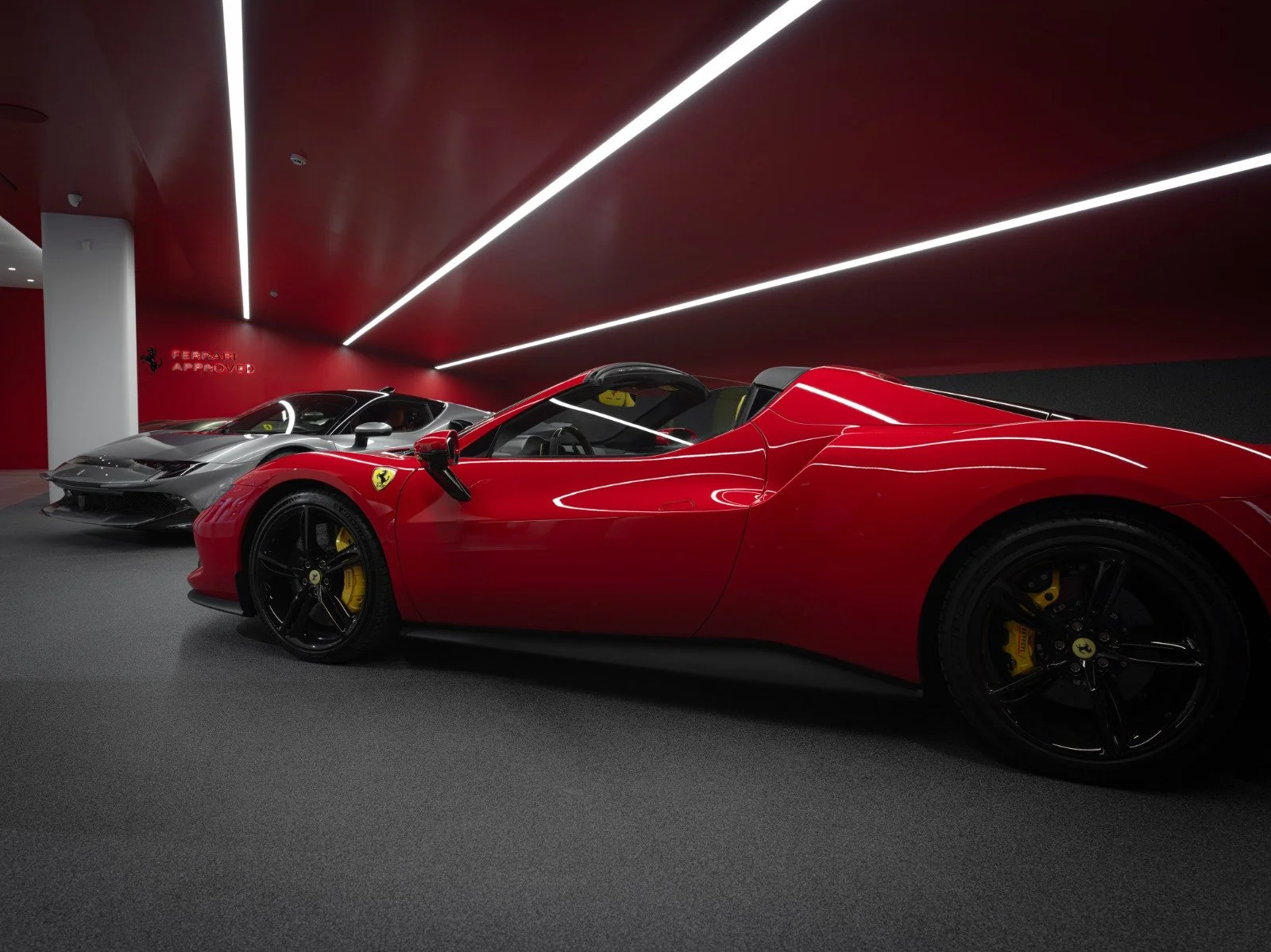 Two luxury sports cars in a showroom, one red Ferrari and one silver Ferrari, with a red and black interior, and a Ferrari approved sign on the wall.