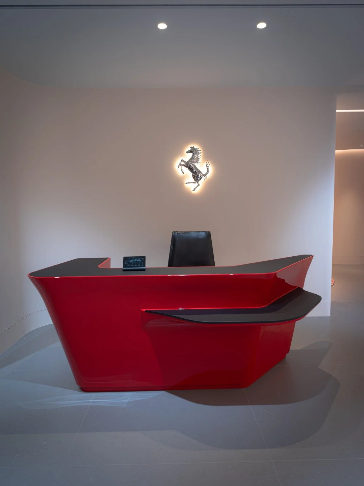 Red and black modern Ferrari office reception desk with a black leather chair behind it, and a Ferrari prancing horse logo illuminated on the wall above.