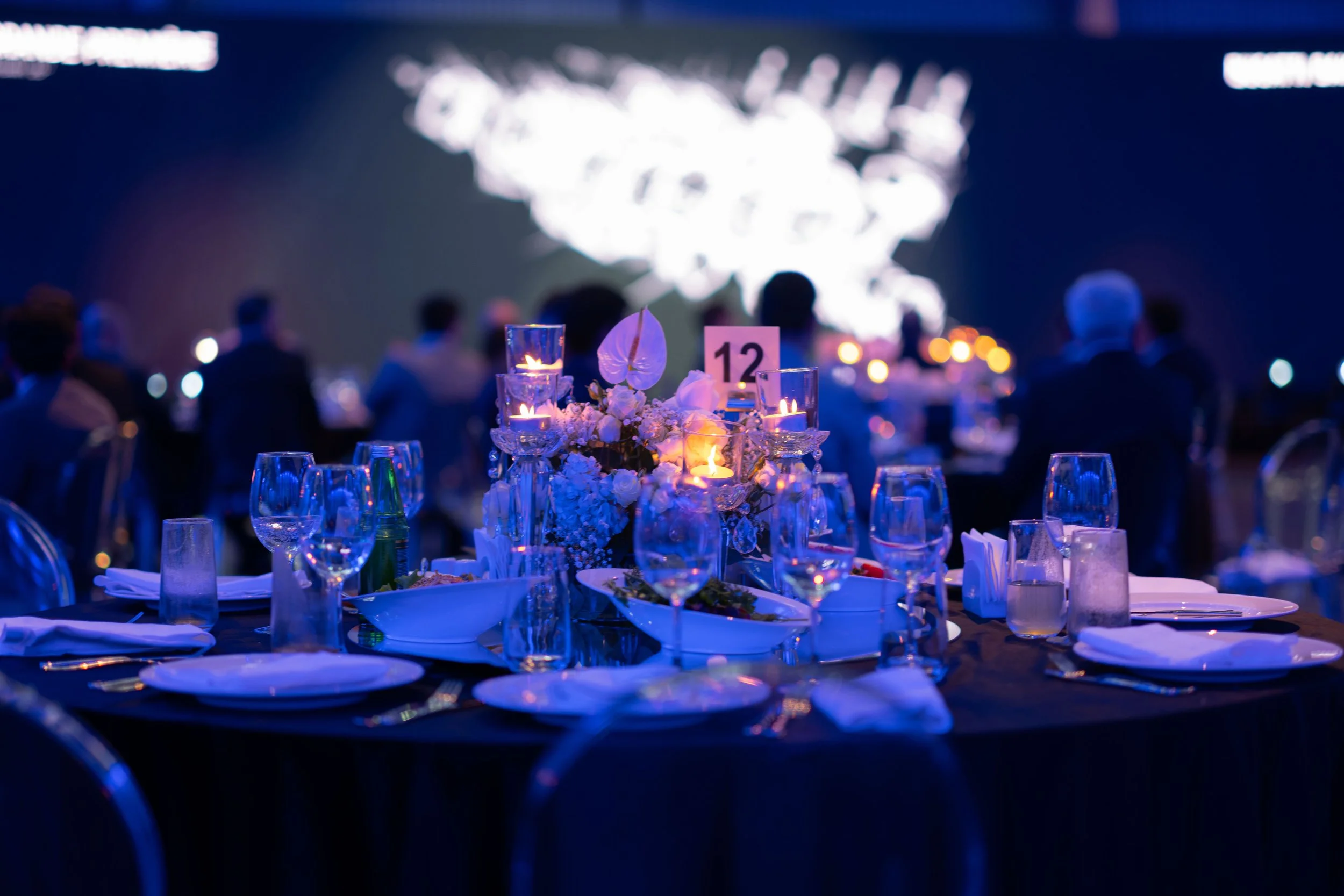 A formal dining table set for an event, decorated with lit candles, floral arrangements, wine glasses, and tableware, with people seated in the background at a dark indoor venue.