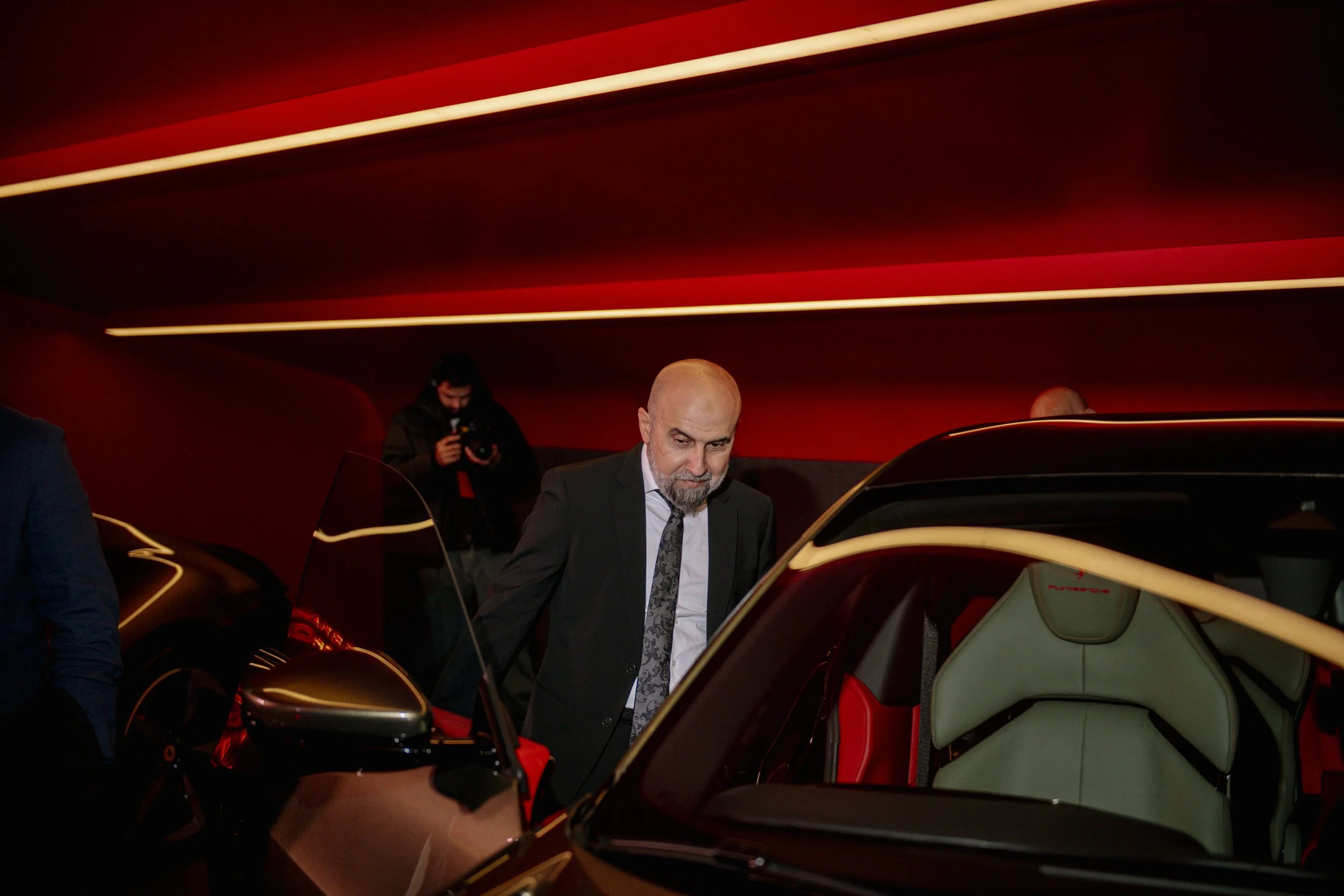 A man in a black suit and patterned tie looking at a car with a green interior, in a red-lit showroom, with a photographer capturing the scene in the background.