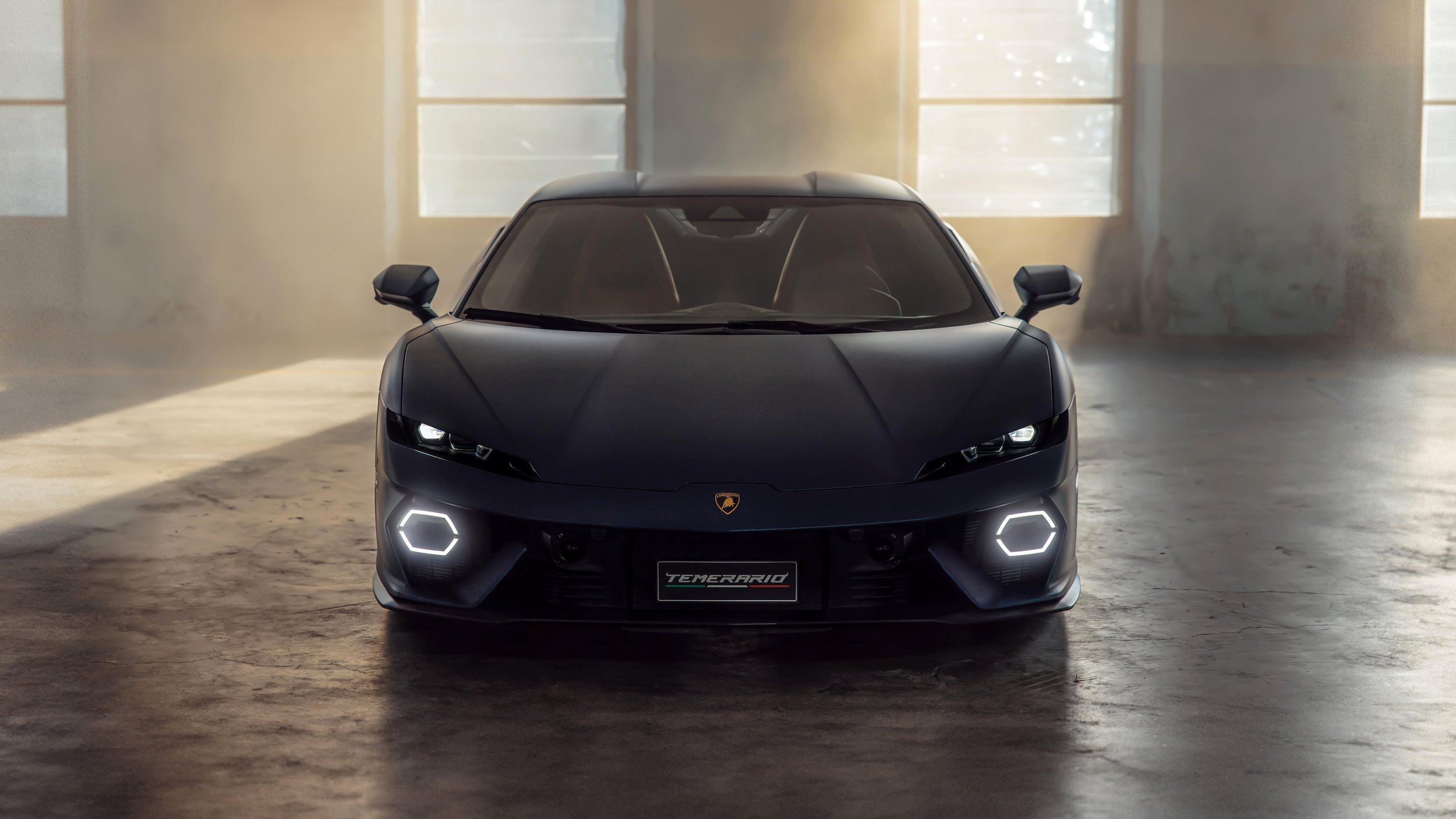 Black sports car with sleek design parked in a dimly lit industrial space, illuminated by natural light from large windows, featuring LED headlights and a Lamborghini emblem on the front