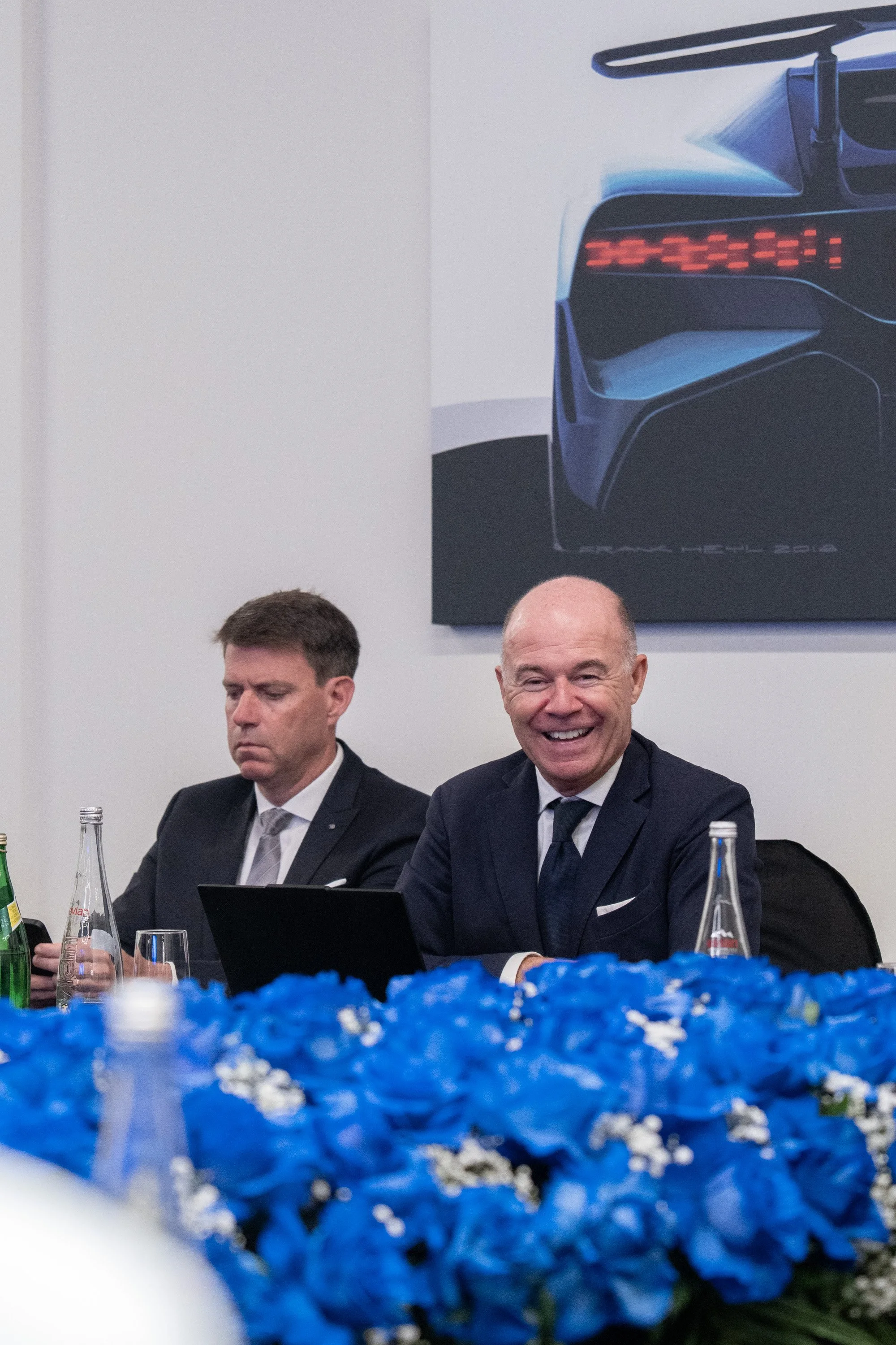 Two men sitting at a table with blue flowers in the foreground. One man is smiling, and the other is looking down. There are bottles of water and glasses on the table. A large poster of a futuristic car is hanging on the wall behind them.