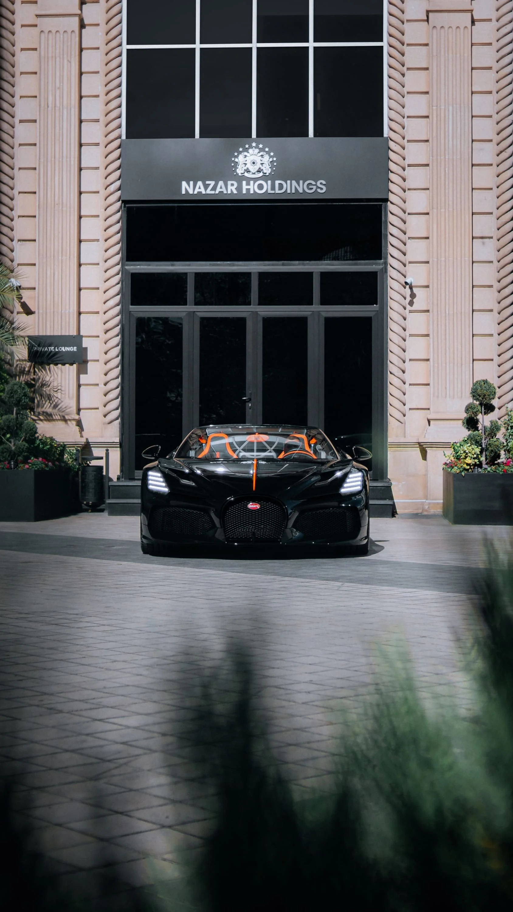 A black luxury sports car parked in front of a building with a sign that reads "NAZAR HOLDINGS." The building has brick walls with vertical columns and large black-framed windows. There are potted plants and a sign for a private lounge to the left.