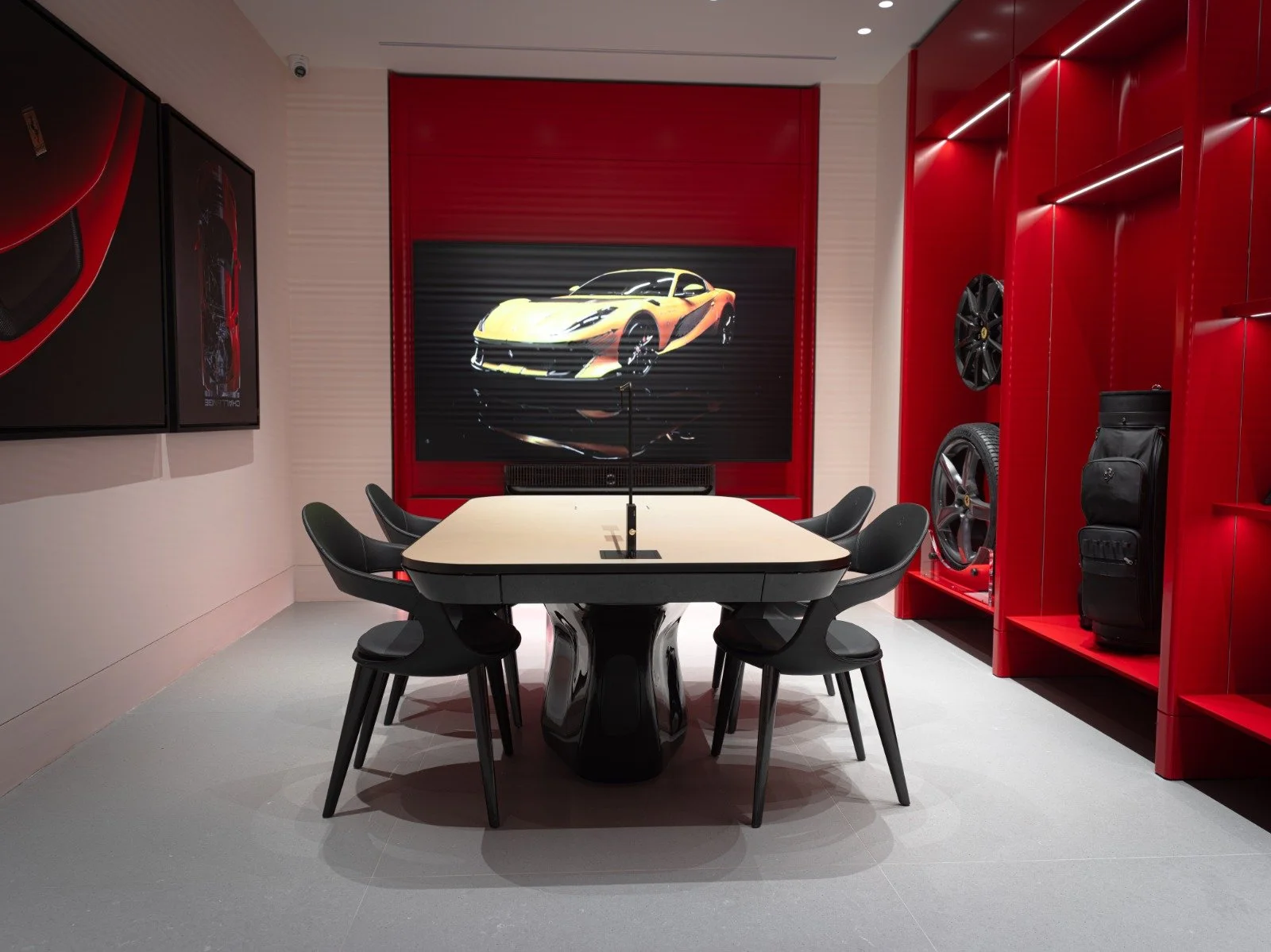 A modern conference room with black chairs around a rectangular table, a large screen displaying a yellow sports car, and red shelves with wheels, tires, a bag, and framed car artwork on the walls.