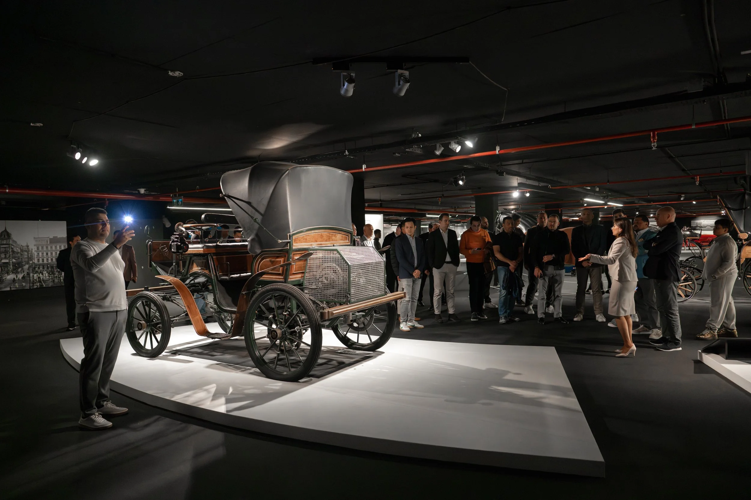 A group of people in a museum gathered around a vintage horse-drawn carriage displayed on a raised platform, with a guide explaining it.