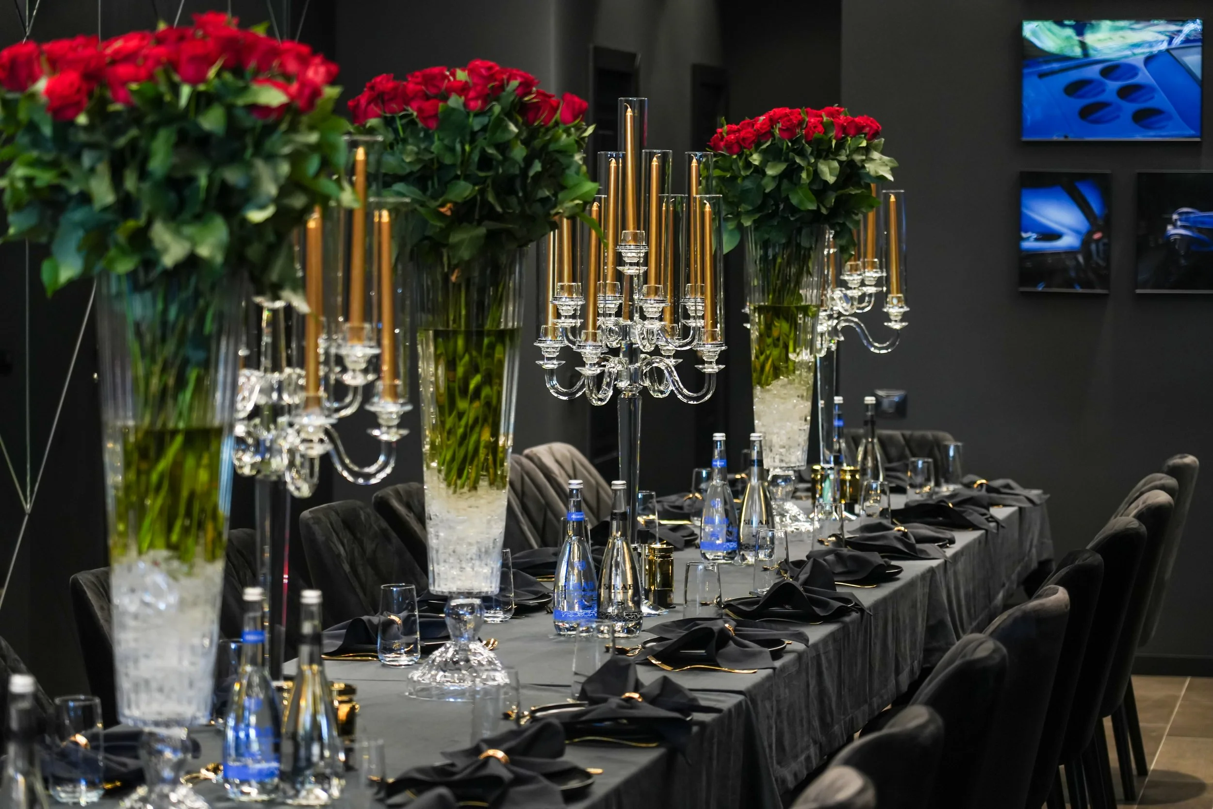 Elegant dining table setup with tall vases of red roses, crystal candelabras with gold candles, black napkins, and water bottles, set against a dark wall with framed artwork.