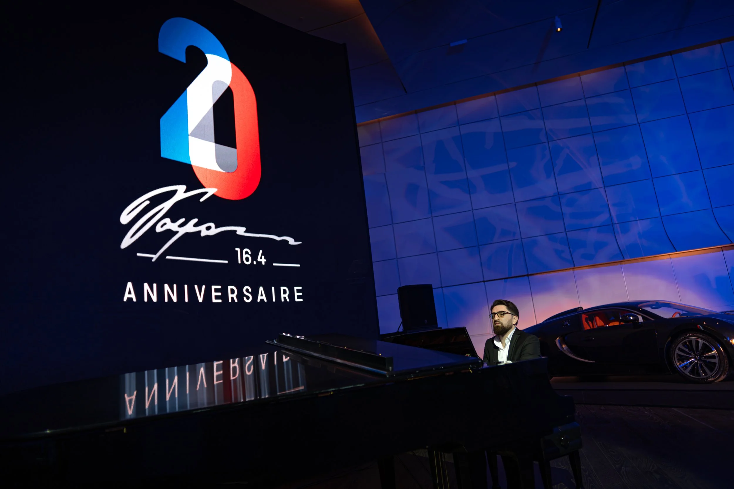 A man playing piano at an event celebrating the 16.4th anniversary of Dior, with a large screen displaying the Dior logo and anniversary text, and a luxury sports car parked nearby.