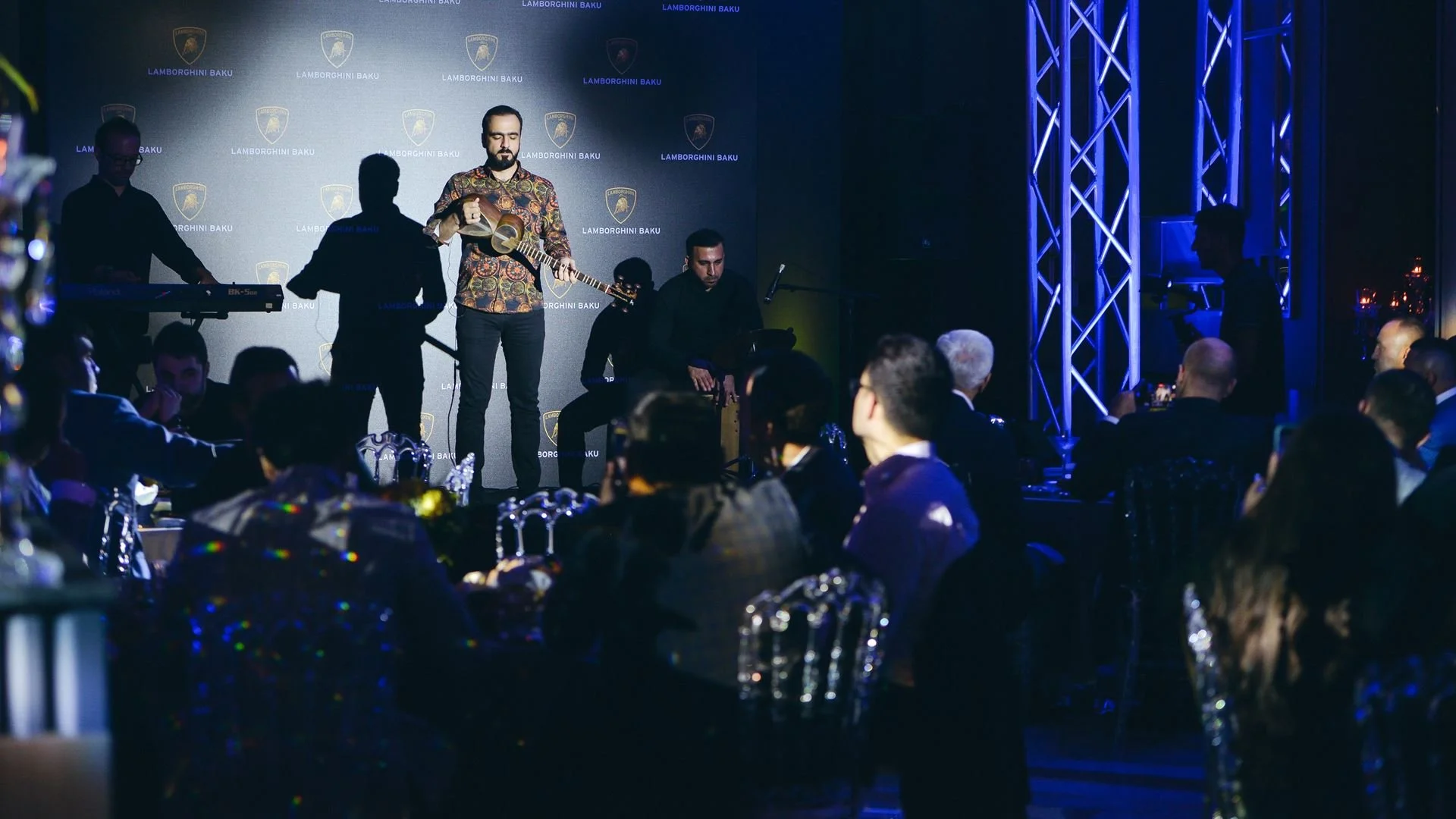 A live musical performance at an event with the Lamborghini Baku logo visible in the background. The performers include a man playing an acoustic guitar and others on keyboard and percussion. The audience seated at tables watches the performance.