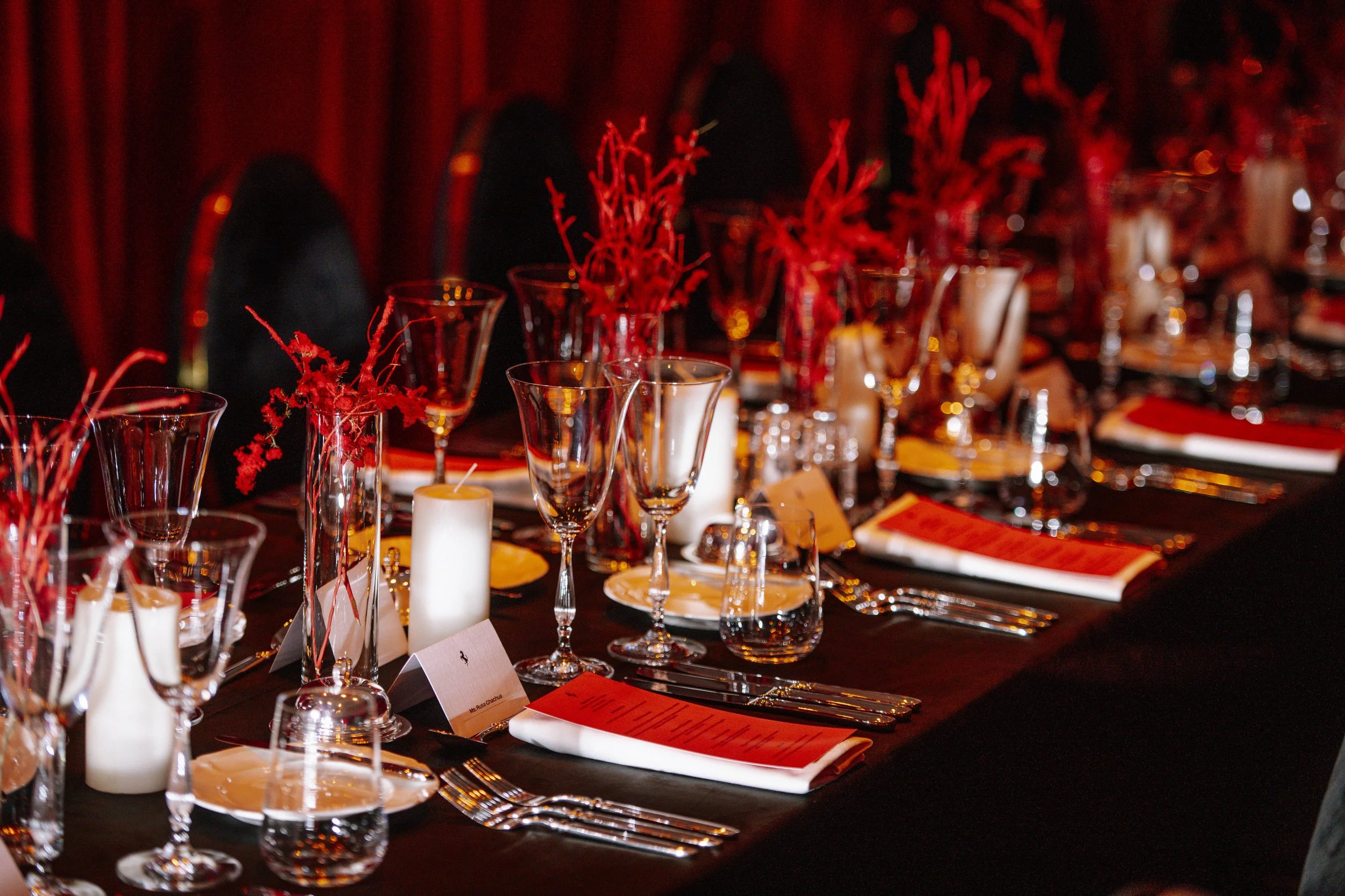 Elegant dining table set with glassware, plates, silverware, candles, and red decorative centerpieces in a dimly lit room.