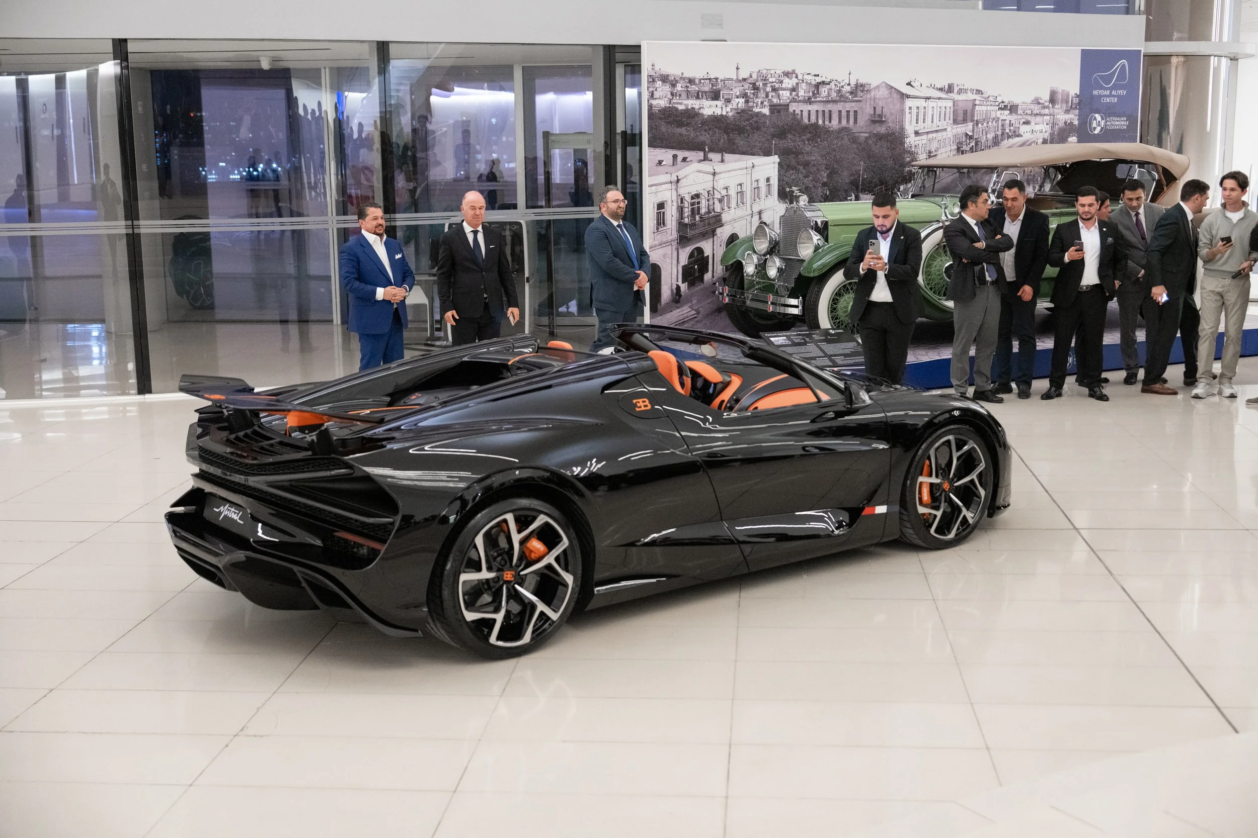 A black Bugatti sports car with orange interior on display at an indoor event, with a group of people in formal attire observing and taking photos in the background.