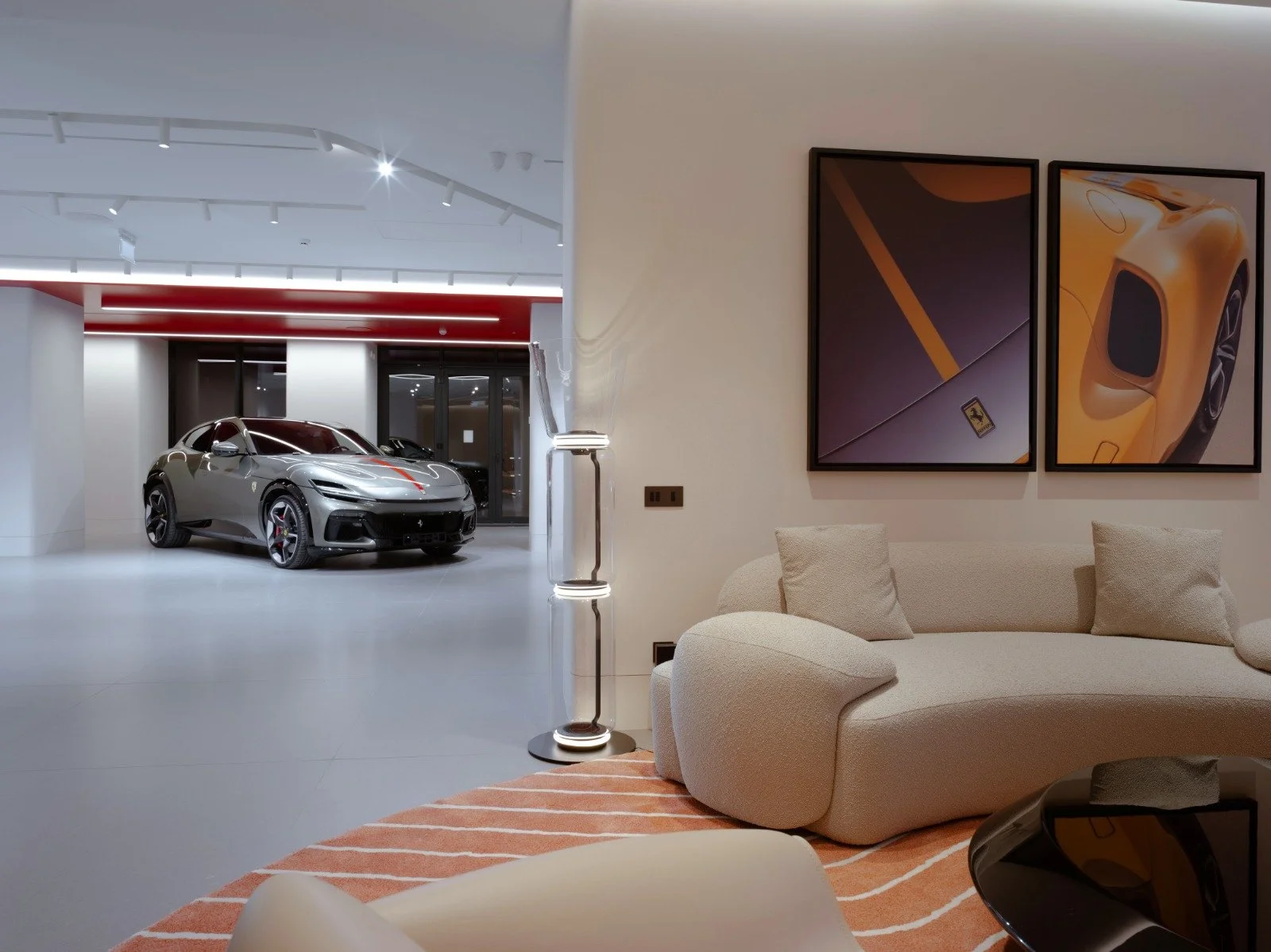 Luxury sports car in a modern showroom seen from the living room of a stylish apartment, which features a beige sofa, framed pictures of a yellow sports car, and a floor lamp.