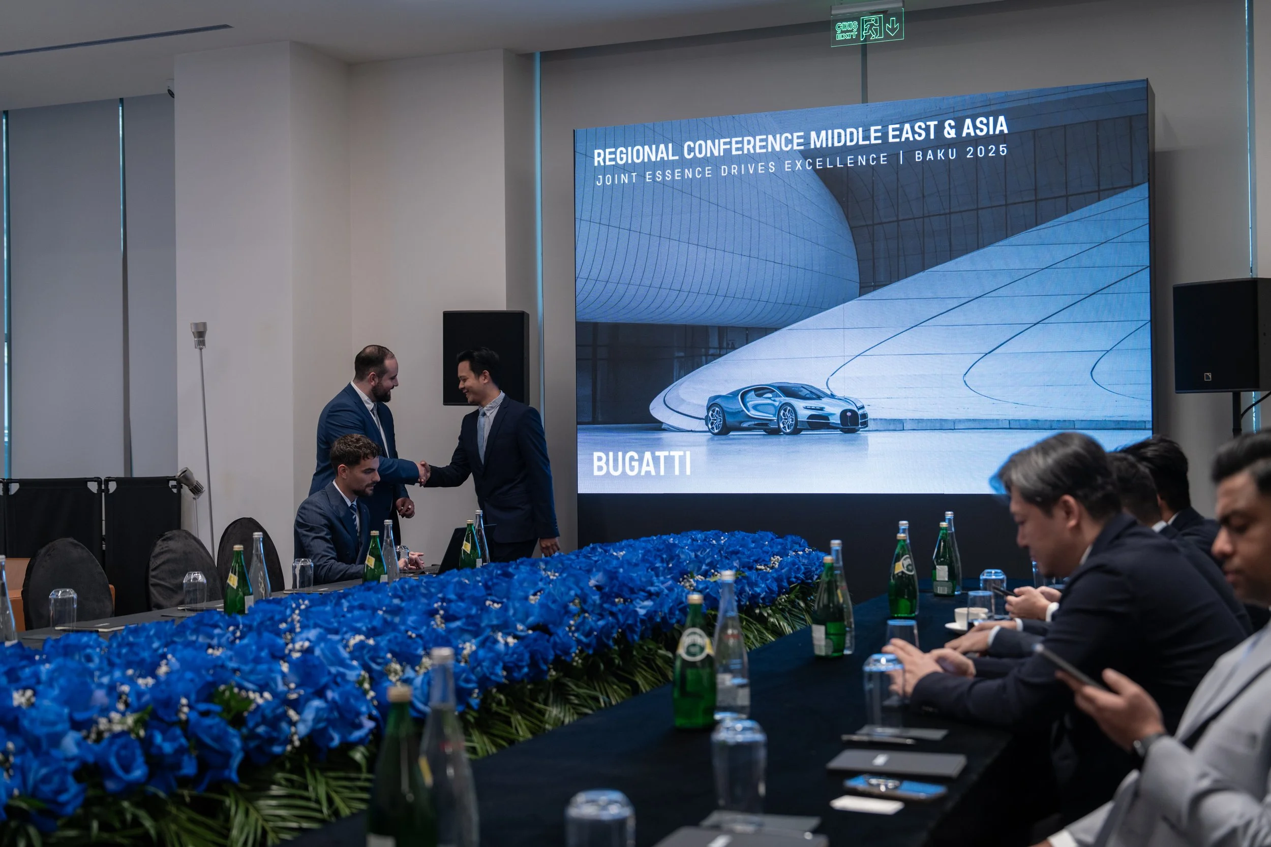 Business meeting at a conference with a large screen displaying 'Regional Conference Middle East & Asia, Joint Essence Drives Excellence, Baku 2025, Bugatti.' Two men are shaking hands, one seated and two others are sitting at a long table decorated with blue flowers, with bottles of water and glasses.