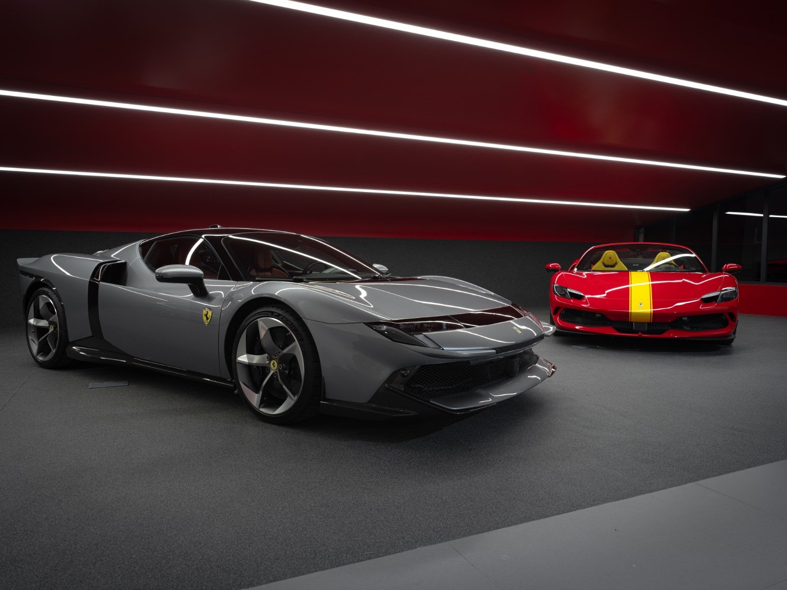 Two luxury Ferrari sports cars, one gray and one red with a yellow stripe, in a modern, illuminated showroom.