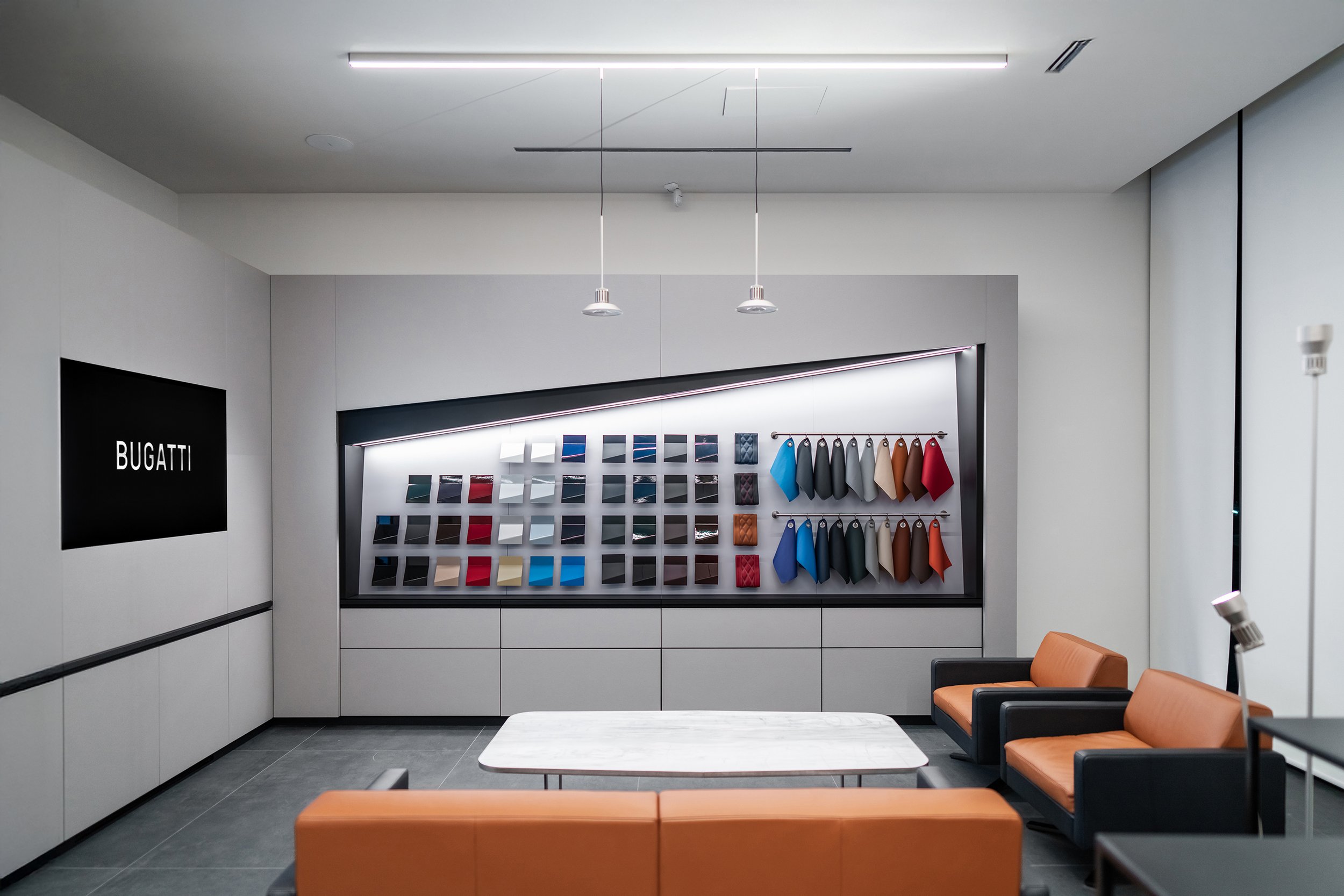 Modern Bugatti showroom with fabric swatches and color samples on a display wall, brown and black leather sofas, and a white marble table.