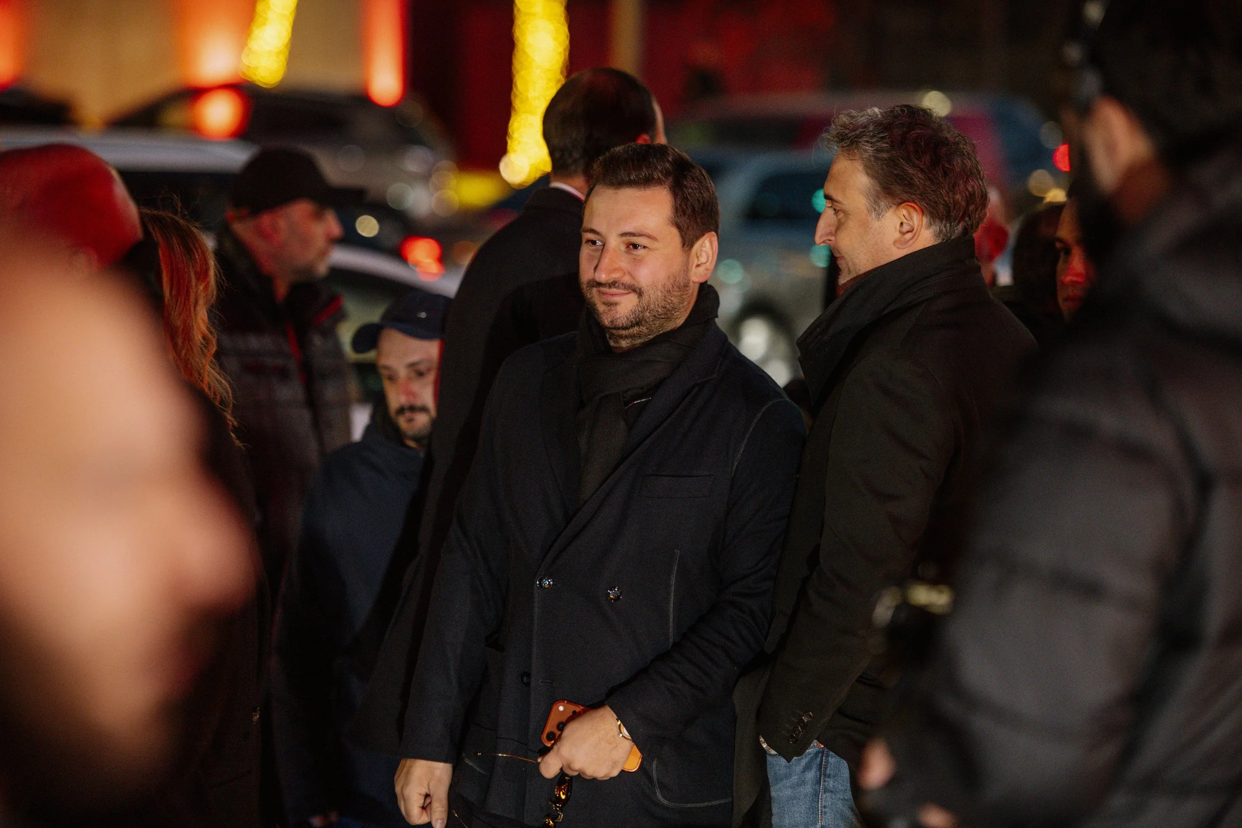 Group of people standing outdoors at night, talking and socializing, with cars and colorful lights in the background.