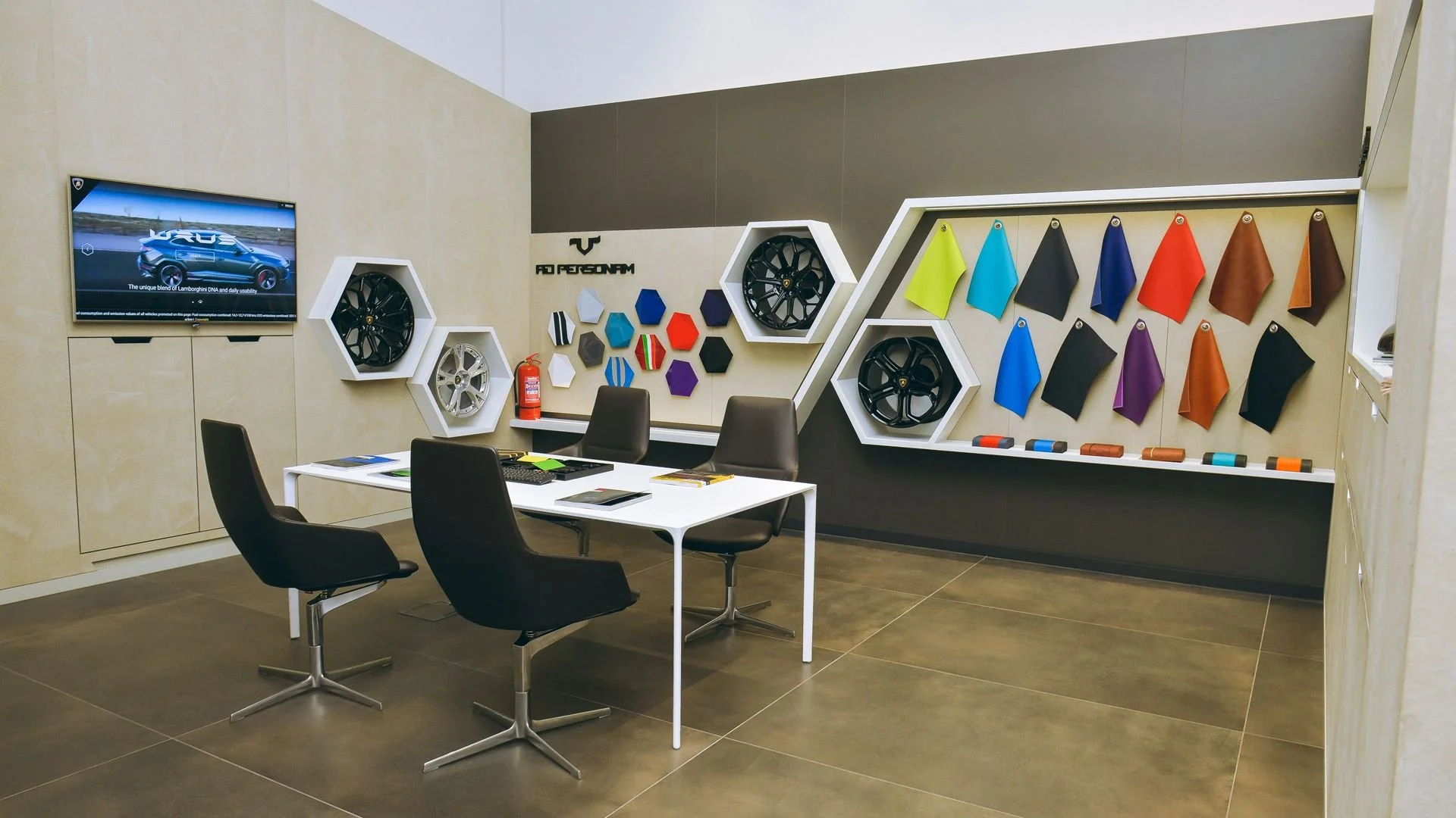 Car dealership or customization shop showroom with a wall display of car wheels and color samples, a TV showing a car, and a white table with four chairs.