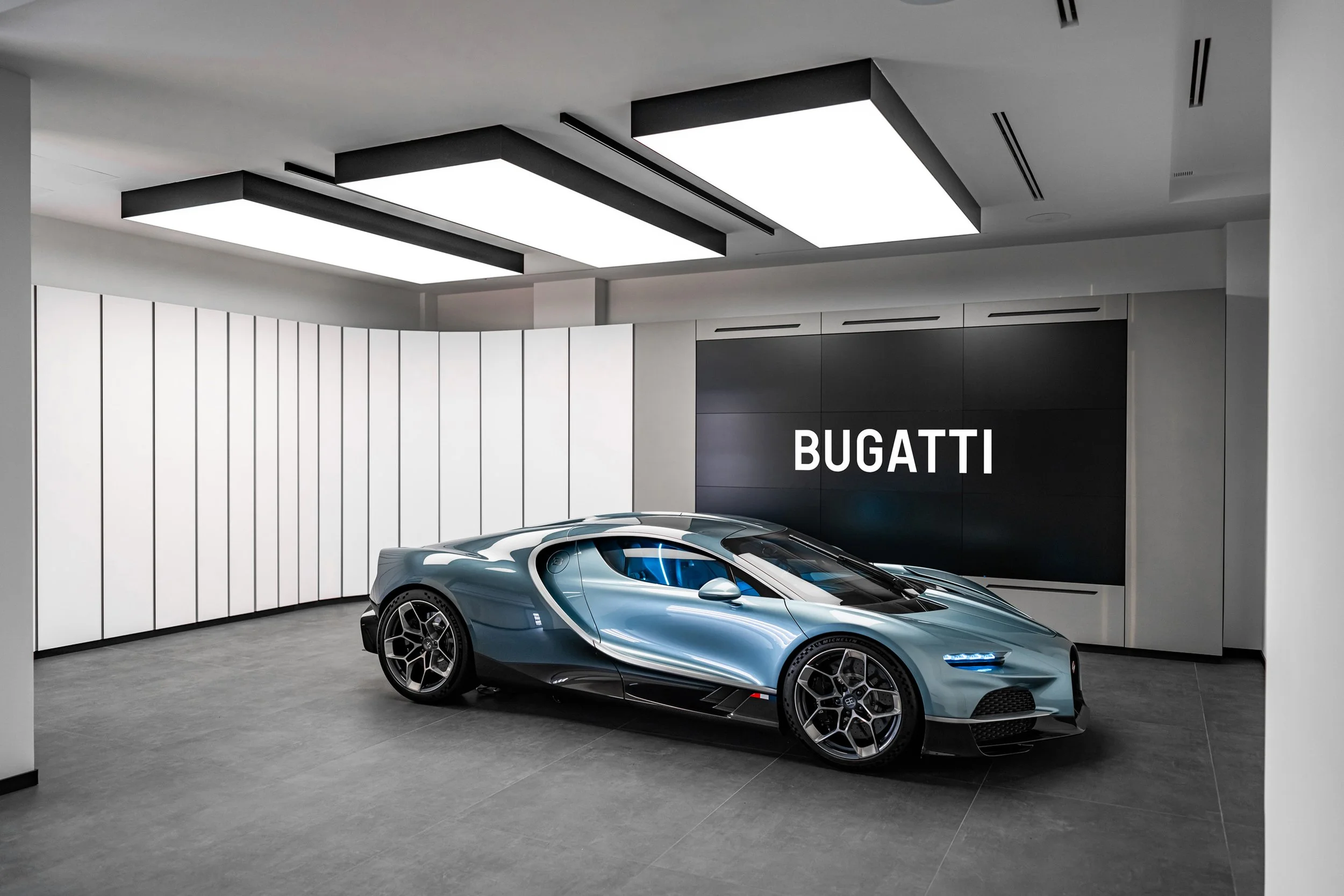 A sleek, blue Bugatti sports car inside a modern showroom with white and black walls, gray flooring, and a large black display screen with the white text 'BUGATTI'.