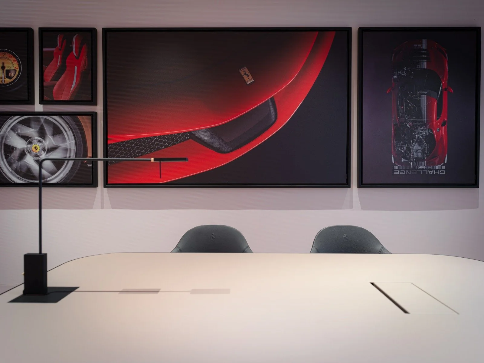 Empty conference table with a modern desk lamp and two chairs in front of three framed automotive posters on the wall, featuring red sports cars and Ferrari logos.