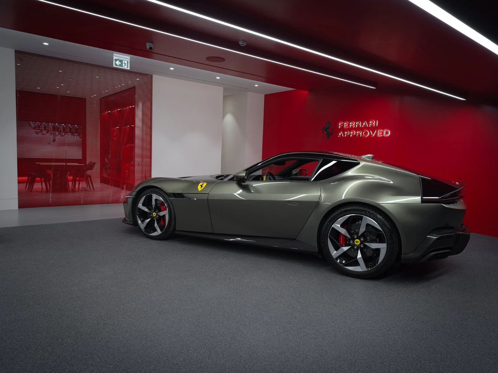 A silver Ferrari sports car inside a showroom with red accents and the Ferrari logo with the words "Ferrari Approved" on a red wall.