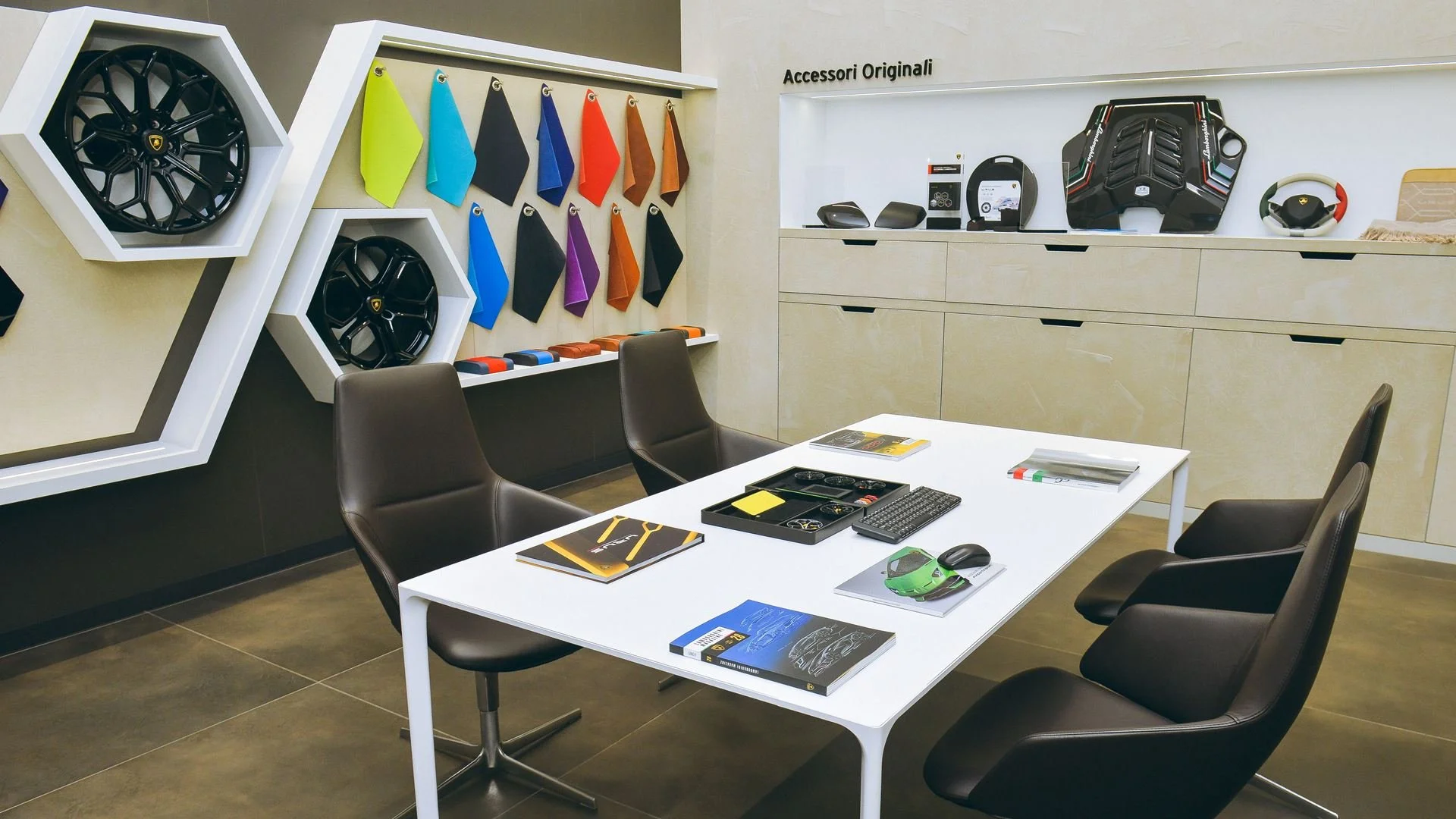 A modern car dealership meeting room with a white table, four black chairs, and a wall display of car accessories including wheels, seat covers, and car parts, with a cabinet and decorative items in the background.