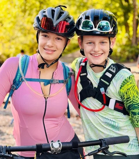Two BEYOND Racing riders in pink and green cycling kits smiling together on the trail.