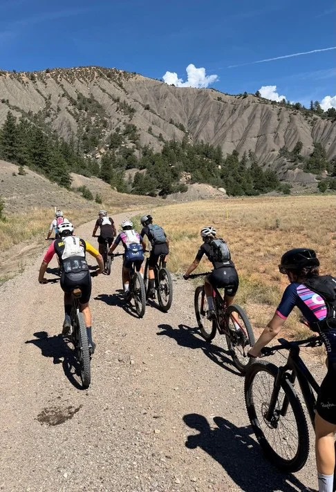 Line of BEYOND Racing mountain bikers riding single file up a gravel trail below a tan rock cliff formation near Durango.