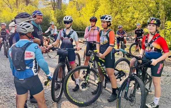 BEYOND Racing riders gathered with their bikes on a gravel trail while a coach in a blue helmet talks with the group.