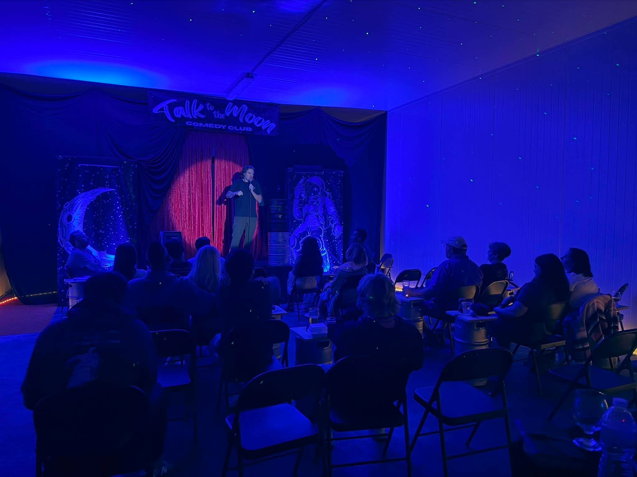 Stage with curtain and starry ceiling at comedy club, audience seated facing performer holding a microphone.