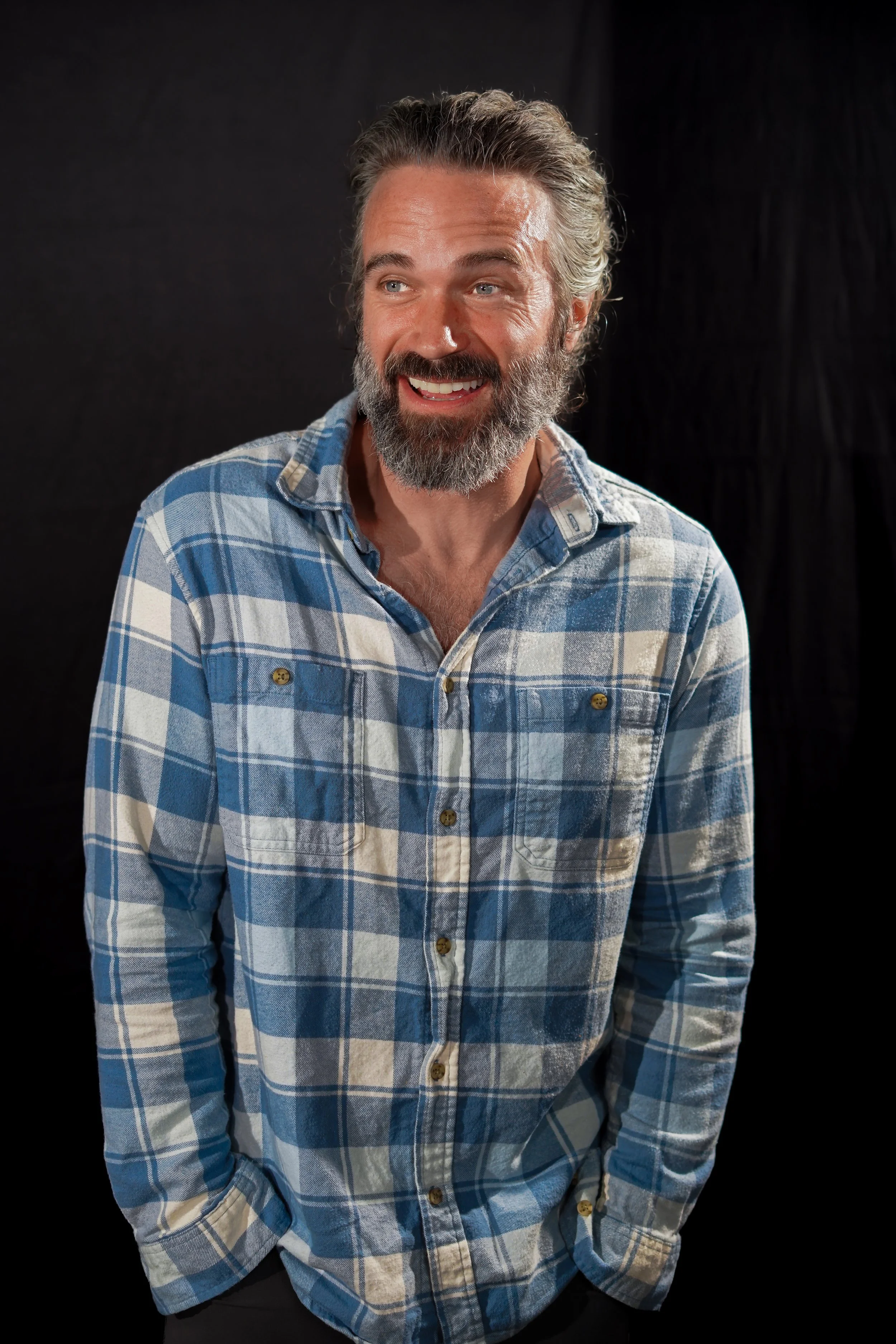 A smiling man with gray hair and beard wearing a blue and white checkered shirt against a black background.