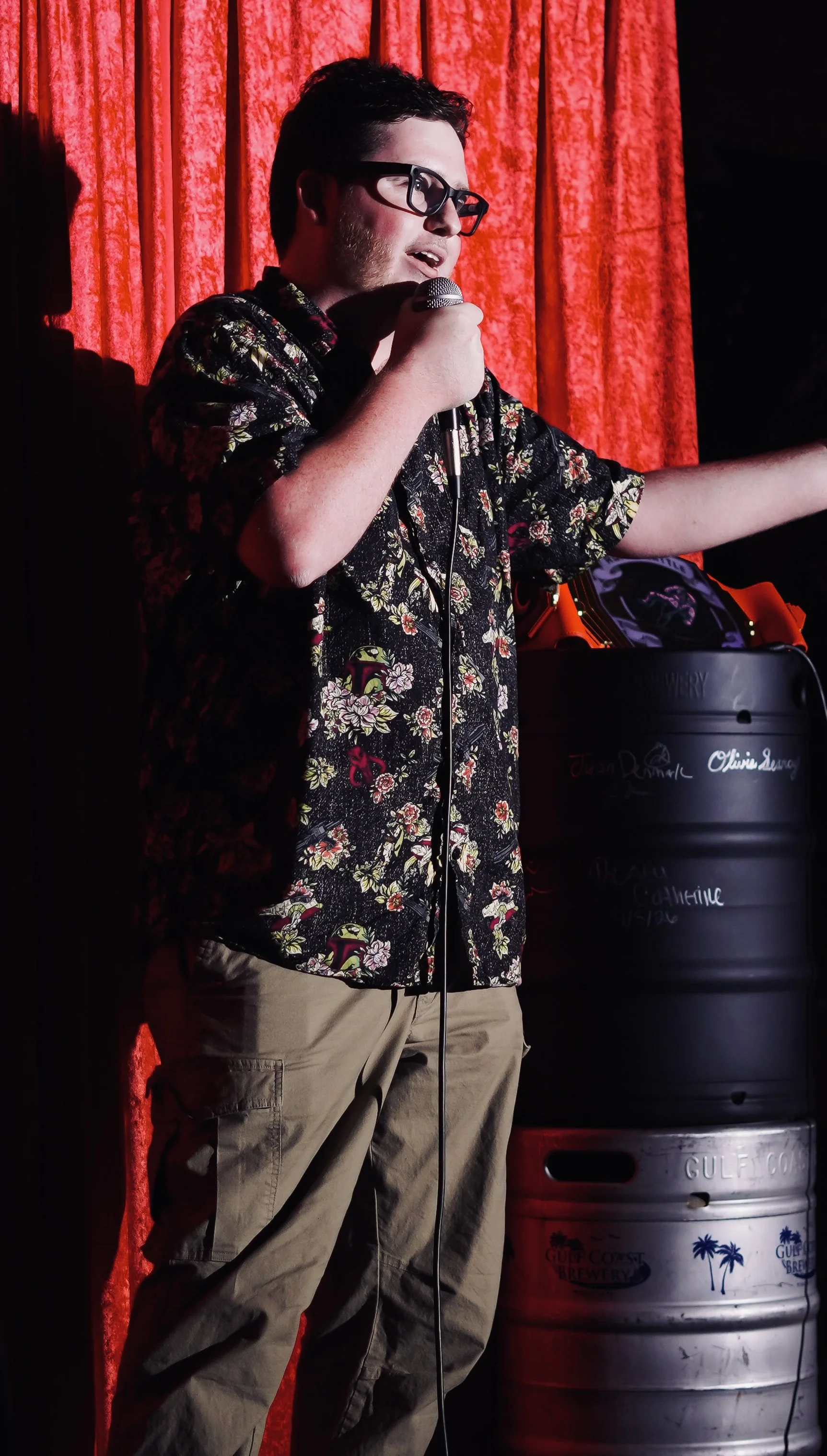 A man wearing glasses and a black floral shirt is holding a microphone and speaking on stage with a red curtain backdrop. There is a metal barrel with sign writing on it in the background.