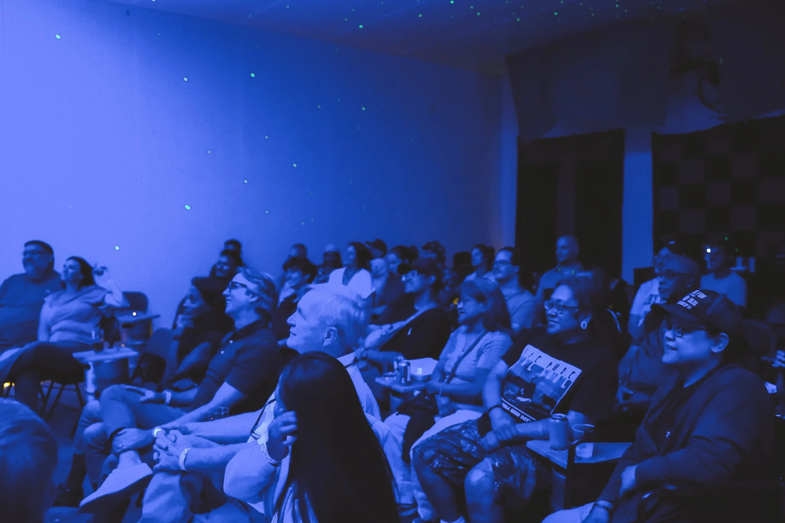 Audience seated in a dark room with a blue tint, watching a presentation or performance. The wall behind has stars or sparkles, and the audience appears engaged.