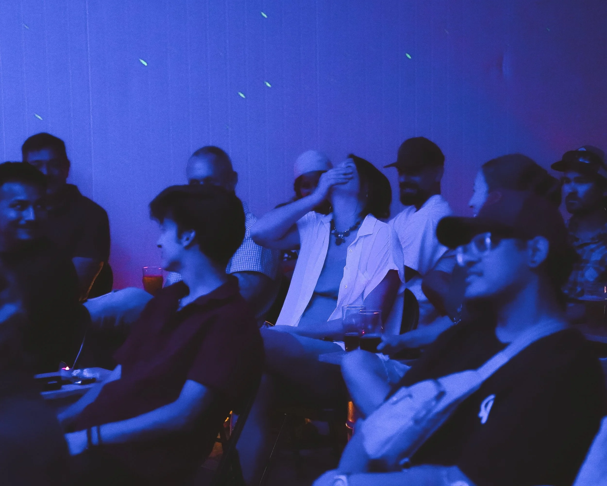 A group of people in a dark room with a blue light, some wearing glasses and hats, and one person in white a shirt, with a woman holding her head.