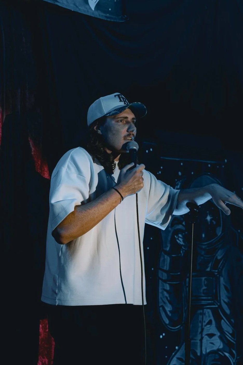 Person with long hair wearing a baseball cap and a white oversized shirt, holding a microphone and speaking, standing in front of a dark space-themed backdrop.